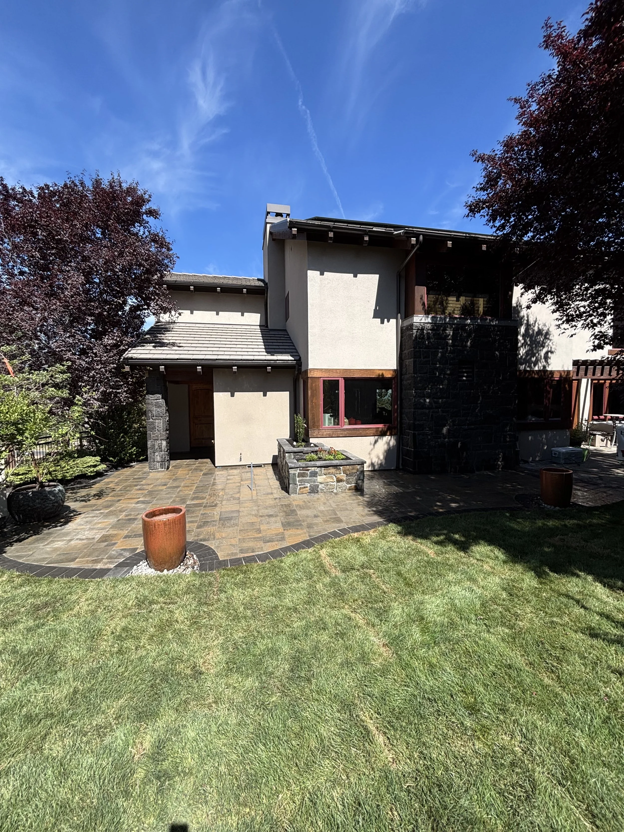A house with a modern design featuring a small balcony and dark stone accents. The yard has a brick patio, a grassy lawn, and decorative plants, with trees on either side and a bright blue sky overhead.