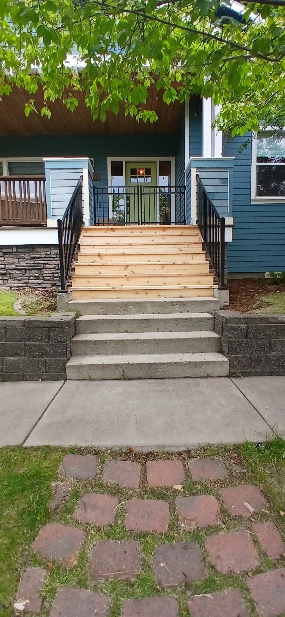 Front steps leading to a green front door with a porch, black railing, and blue house exterior, with red brick pavers and concrete sidewalk in front.
