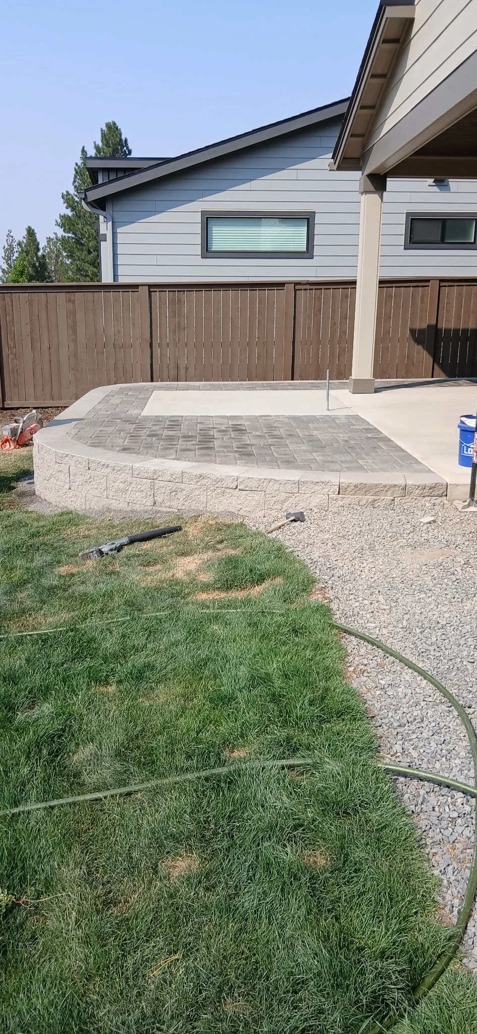 Newly constructed backyard patio with a curved stone wall and paver surface, adjacent to a grassy lawn with construction tools and materials nearby.