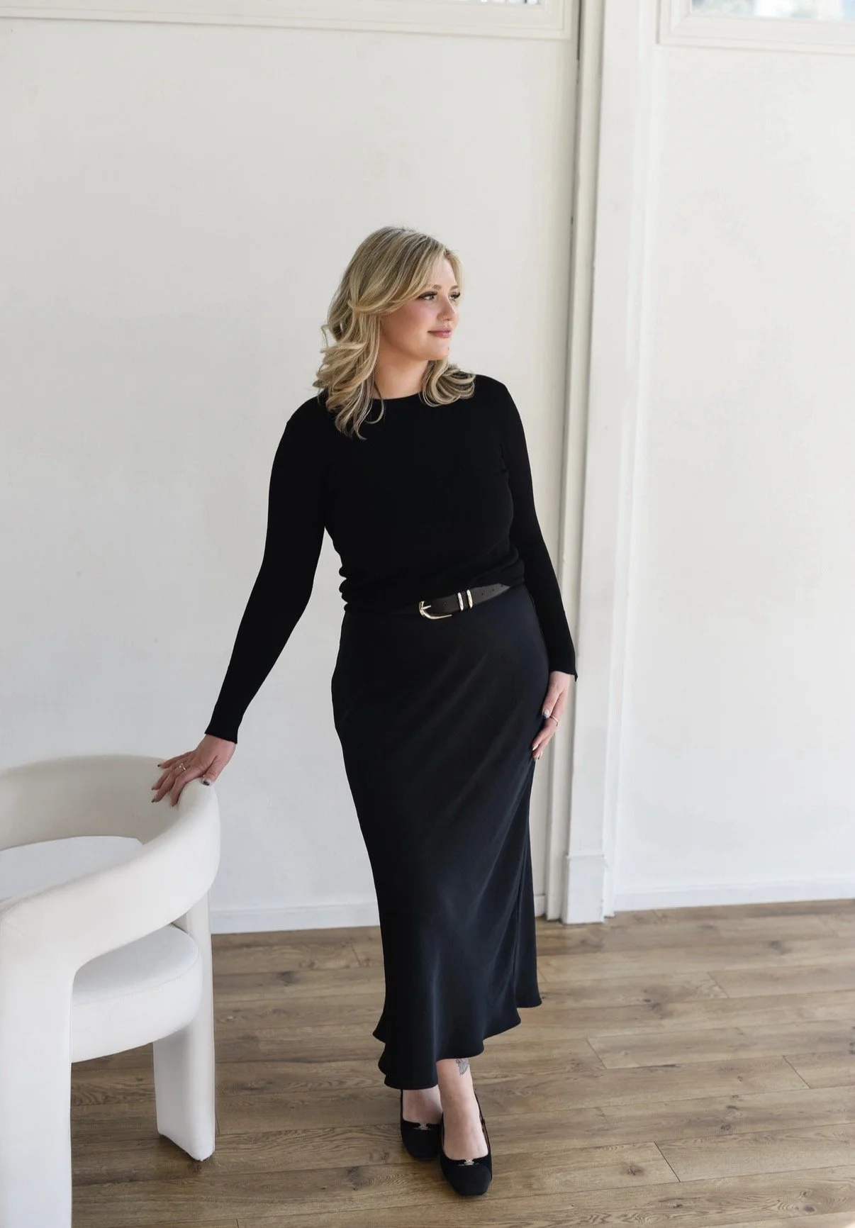 A woman in a black long-sleeve top and long black skirt standing in a minimalist room with white walls and wooden floors, resting her hand on a white chair.