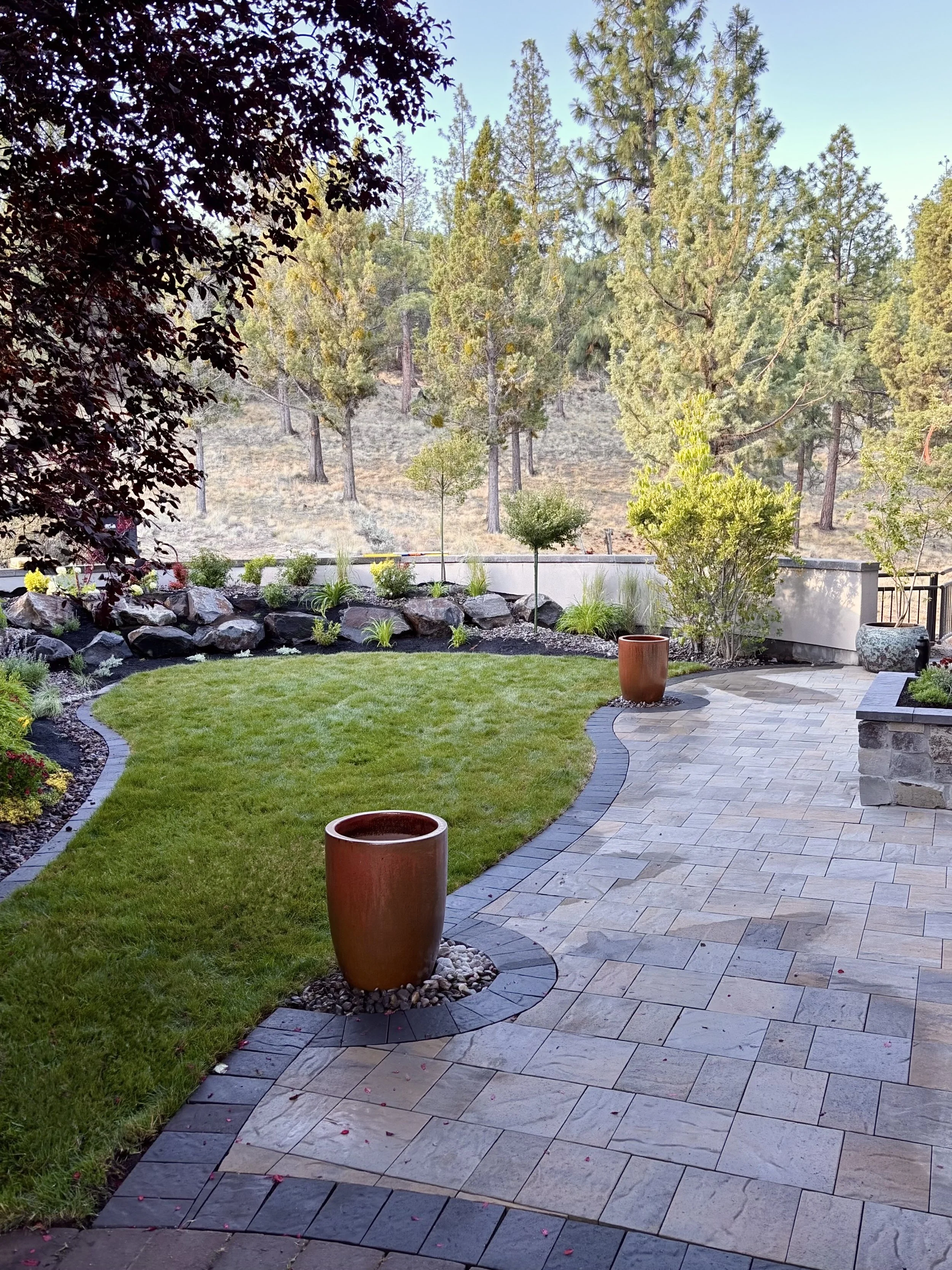 A landscaped backyard with a curved stone and paver patio, green grass, large decorative pots, and a backdrop of trees and a hillside.