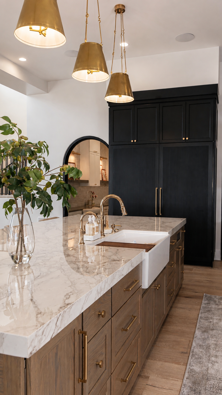A modern kitchen with a marble countertop, a white farmhouse sink, black cabinets with gold handles, a large potted plant, and three brass pendant lights hanging above.