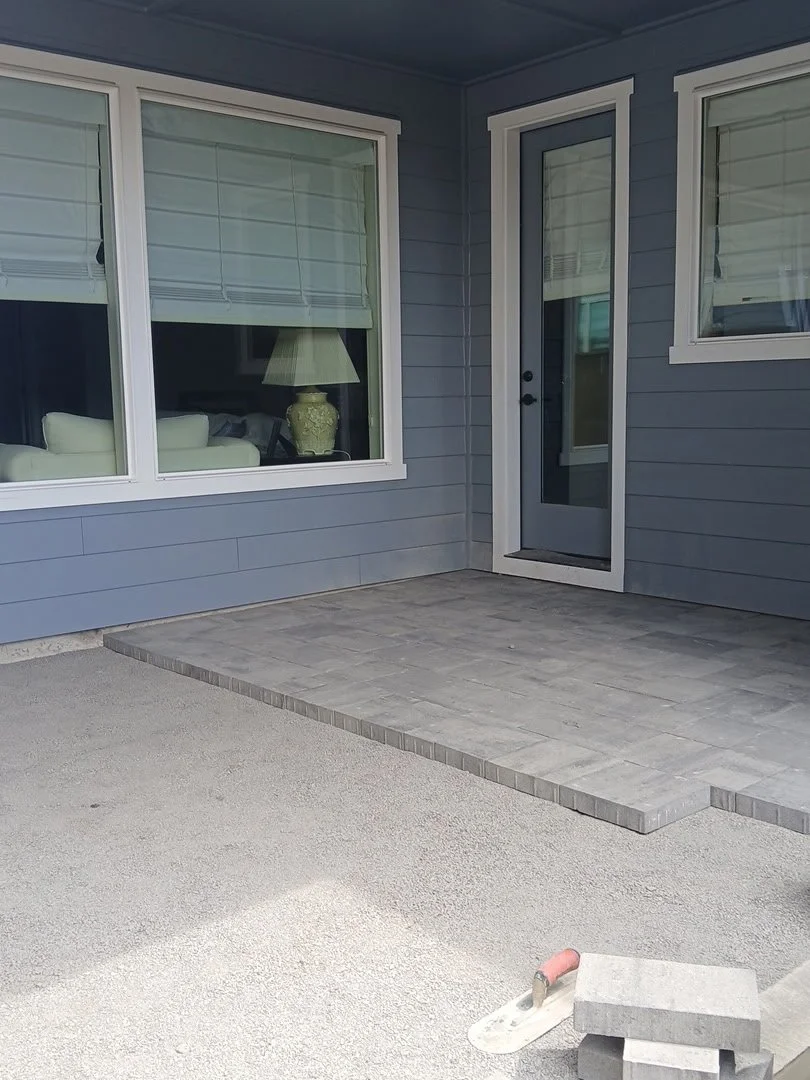 A newly constructed small patio area with gray brick pavers next to a blue house with two large windows and a door, with construction tools and materials in the foreground.