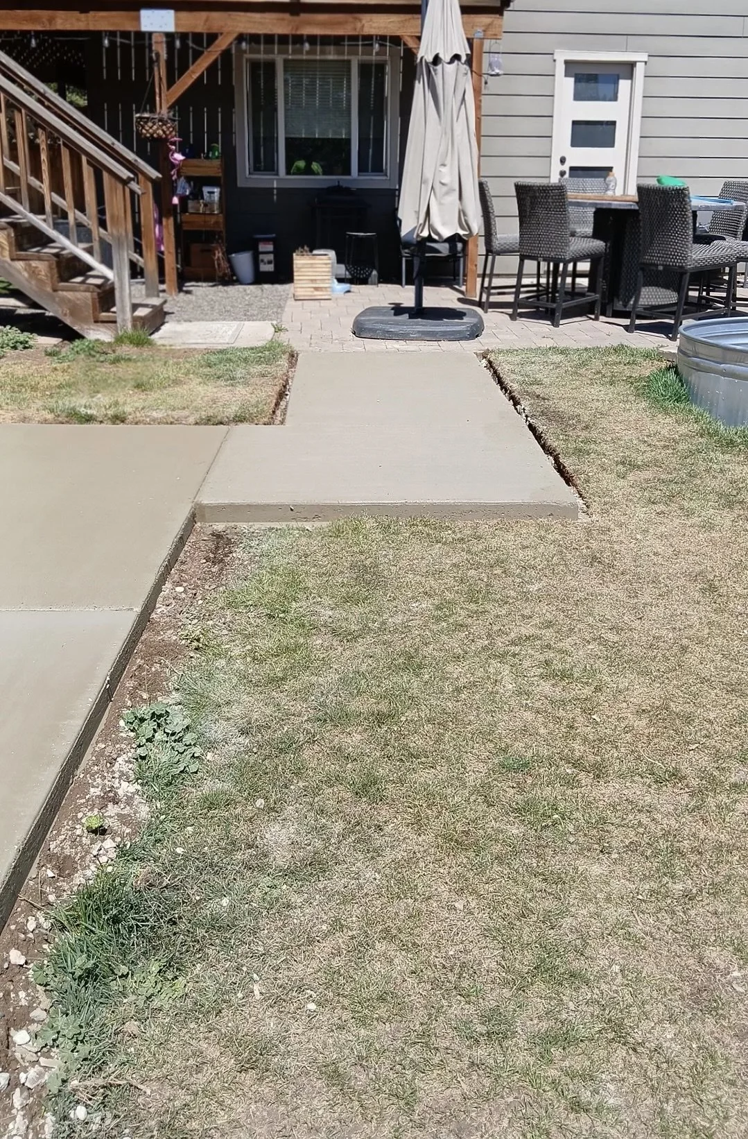 Backyard patio area with concrete walkway, small grassy patches, outdoor dining table with six chairs, umbrella stand without umbrella, stairs leading to house, window, and various outdoor items.