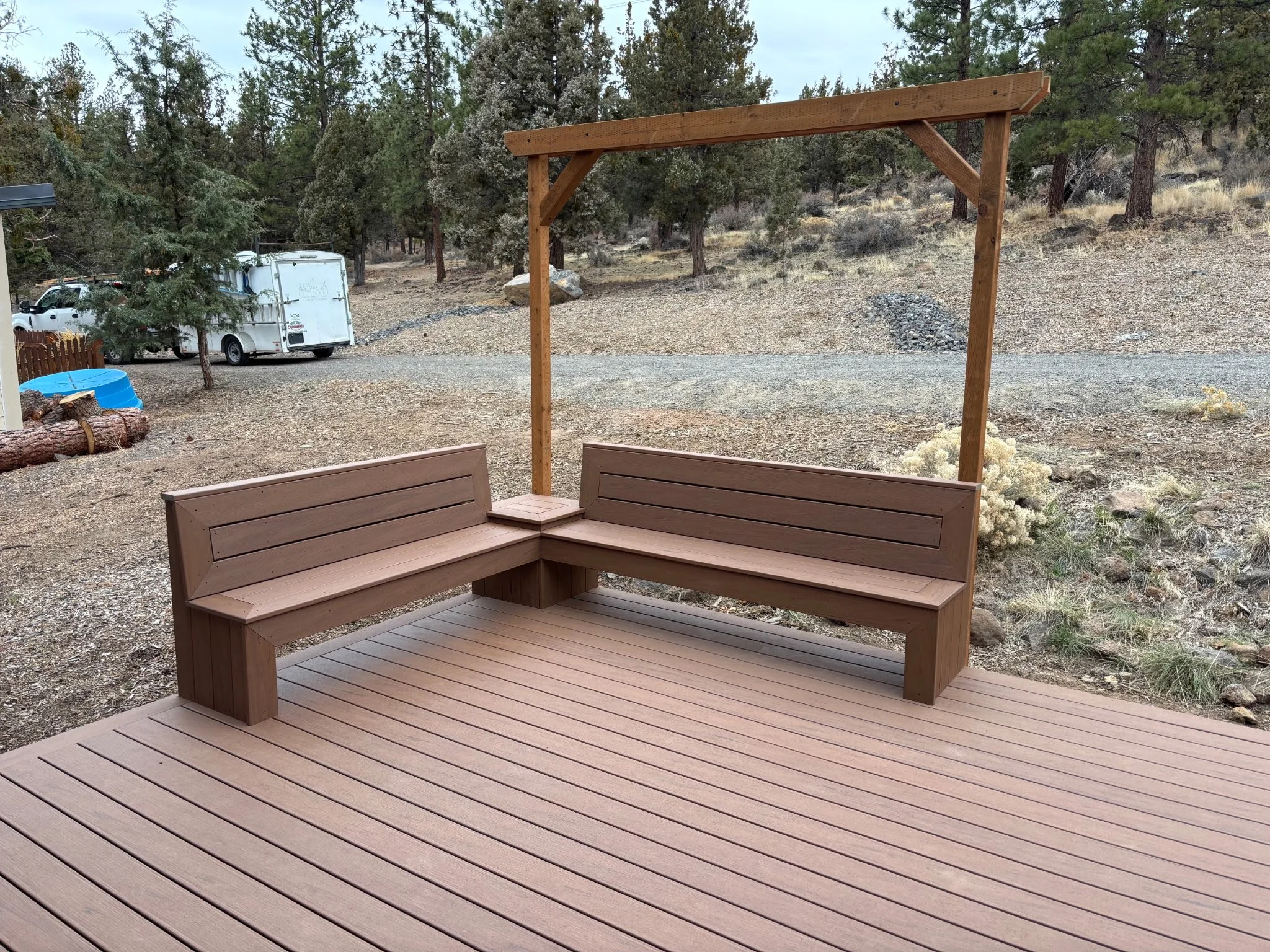 A wooden outdoor living and outdoor seating area with a built-in corner bench and a pergola frame, overlooking a gravel yard with trees and a white trailer in the background.
