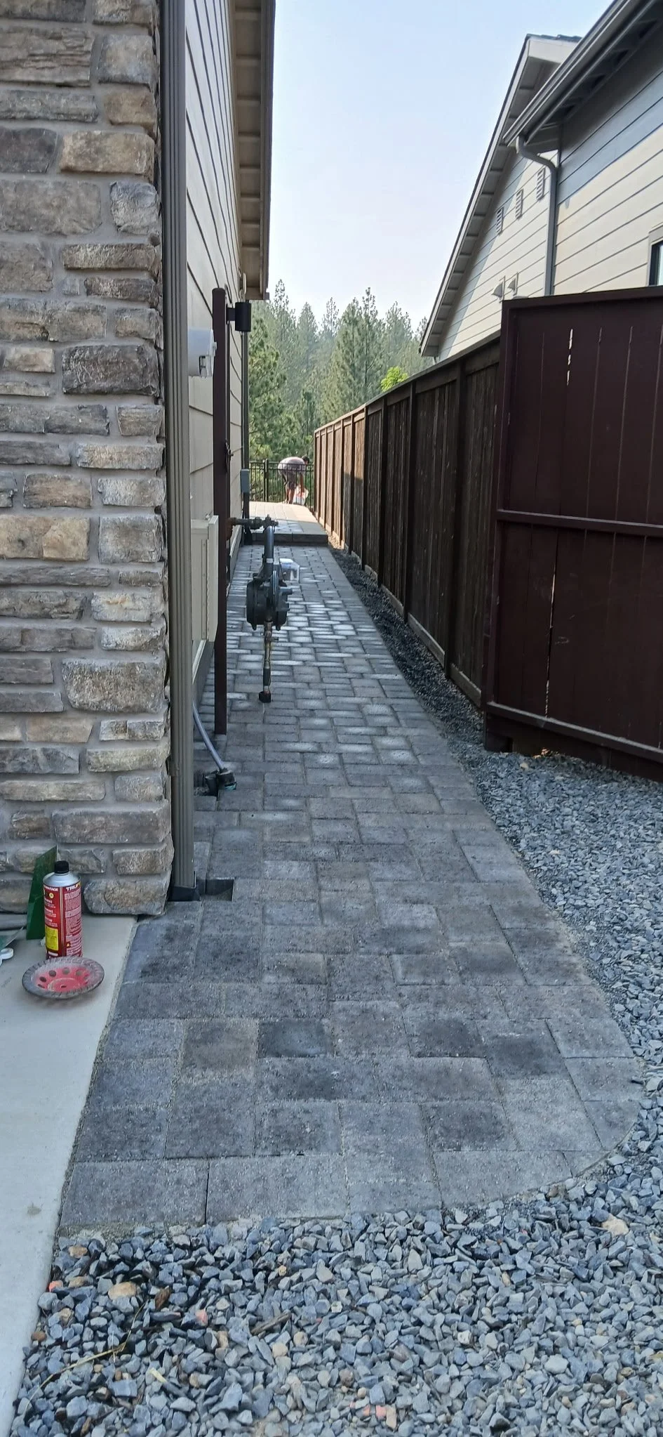 A narrow backyard pathway with brick pavers, flanked by a stone wall on the left and a dark wooden fence on the right, leading toward a wooded area in the background. Retaining wall.