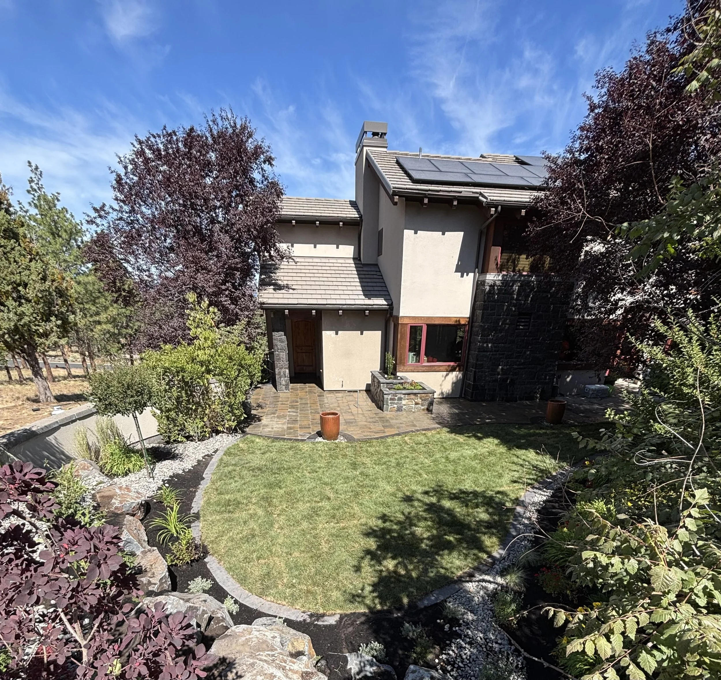 A backyard garden with a house in the background, trees, and a clear blue sky. Paver patio.