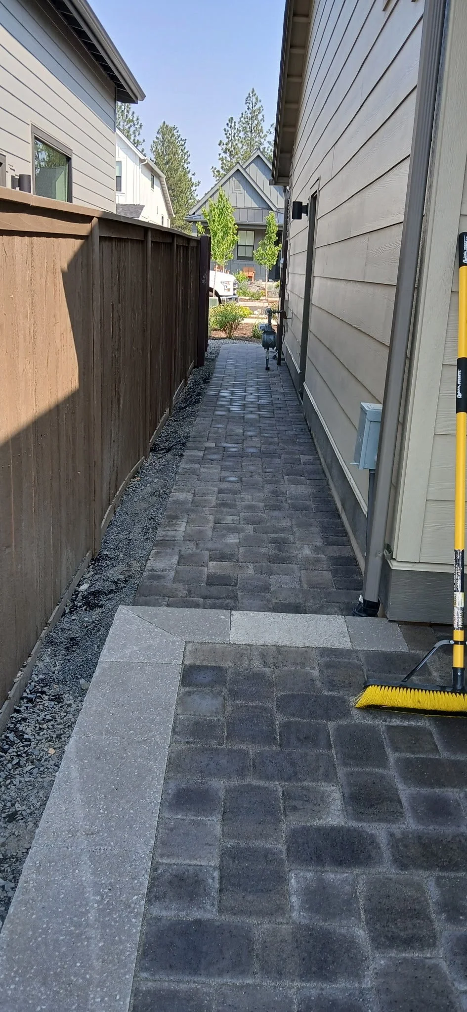 Backyard pathway with brick pavers bordered by concrete, flanked by a wooden fence on the left and the house on the right, with trees and neighboring houses in the background.