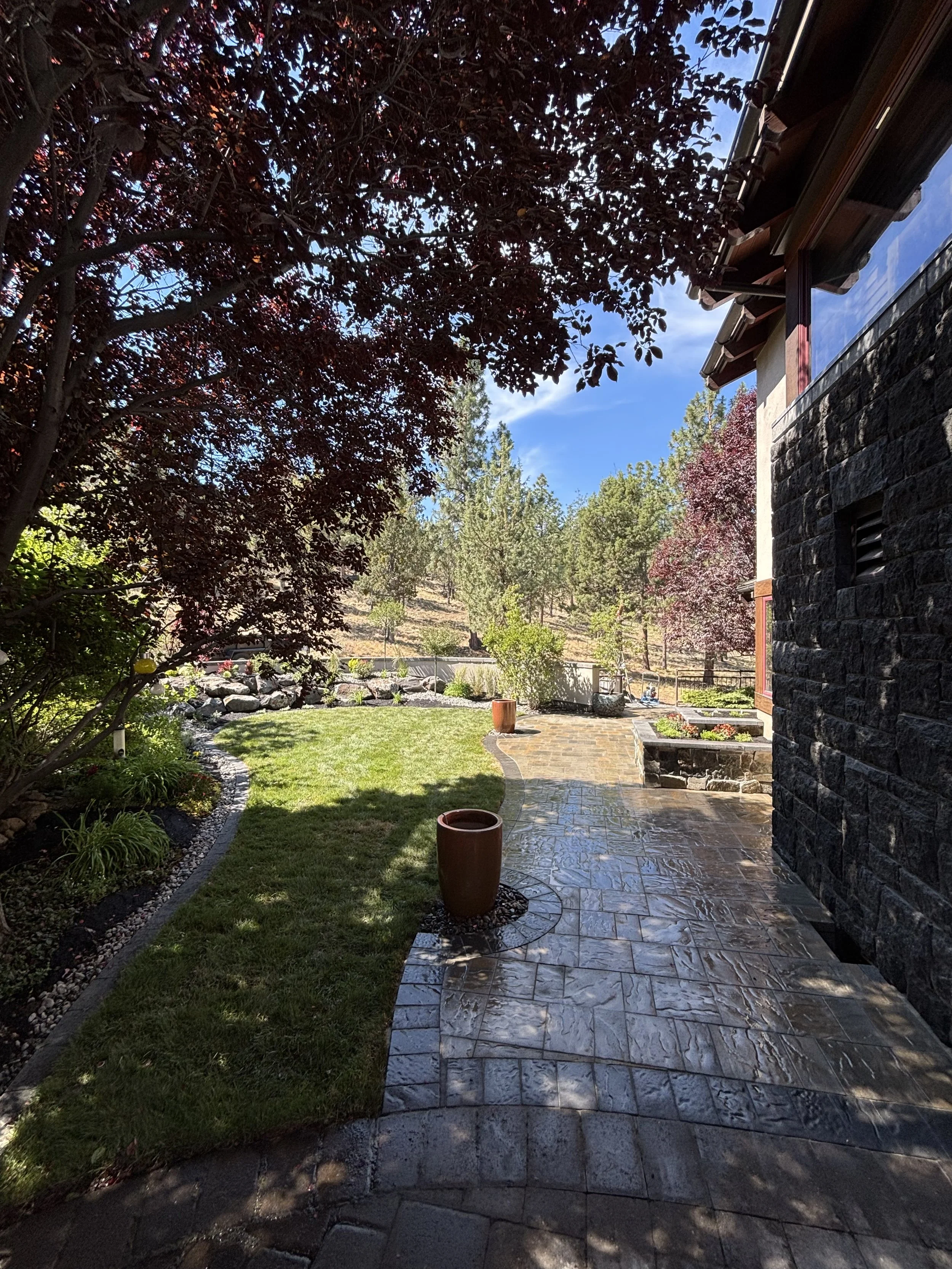 View of a backyard patio with a stone pathway, potted plants, lush green grass, and trees with dark red and green leaves. Part of a house with a stone exterior and large window is visible on the right. The sky is partly cloudy.