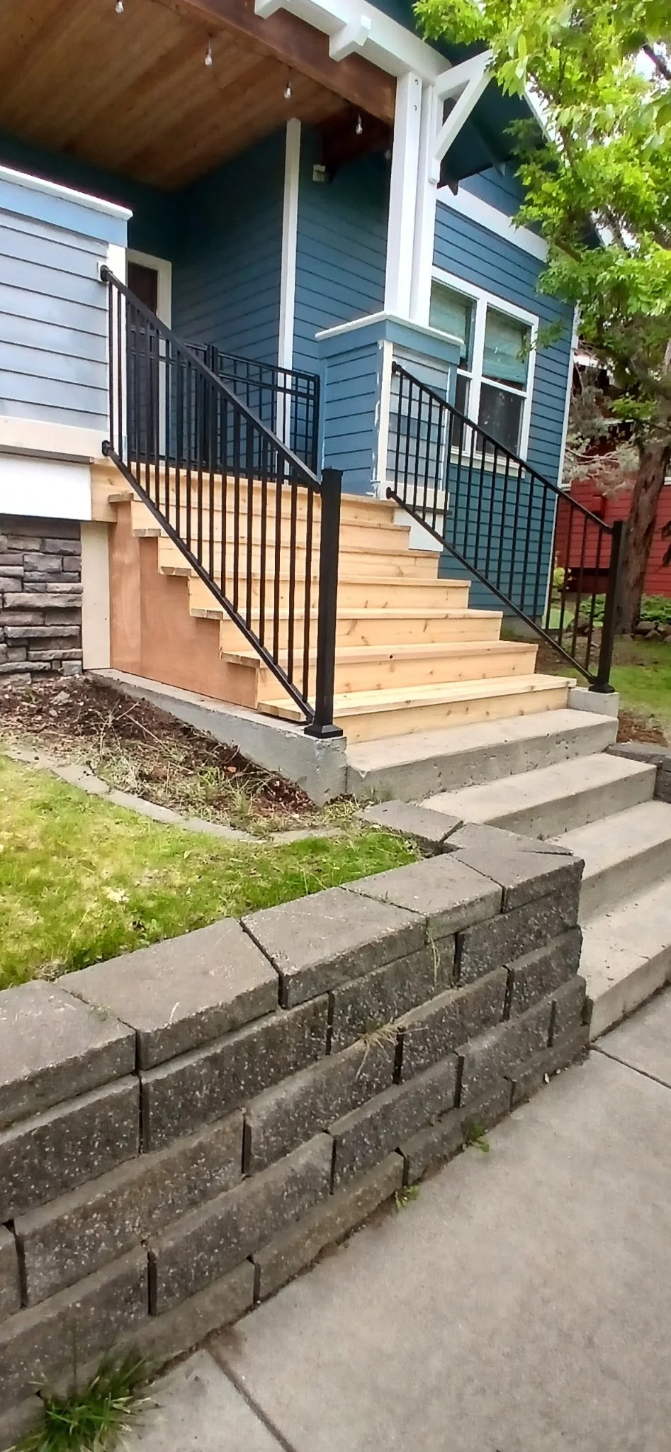A house with blue siding, a wooden porch, and a new set of wooden stairs with black metal railings leading up to the front door. There is a concrete sidewalk and a brick retaining wall in the foreground, with some grass and soil beside the stairs. A tree with green leaves is on the right side of the image.