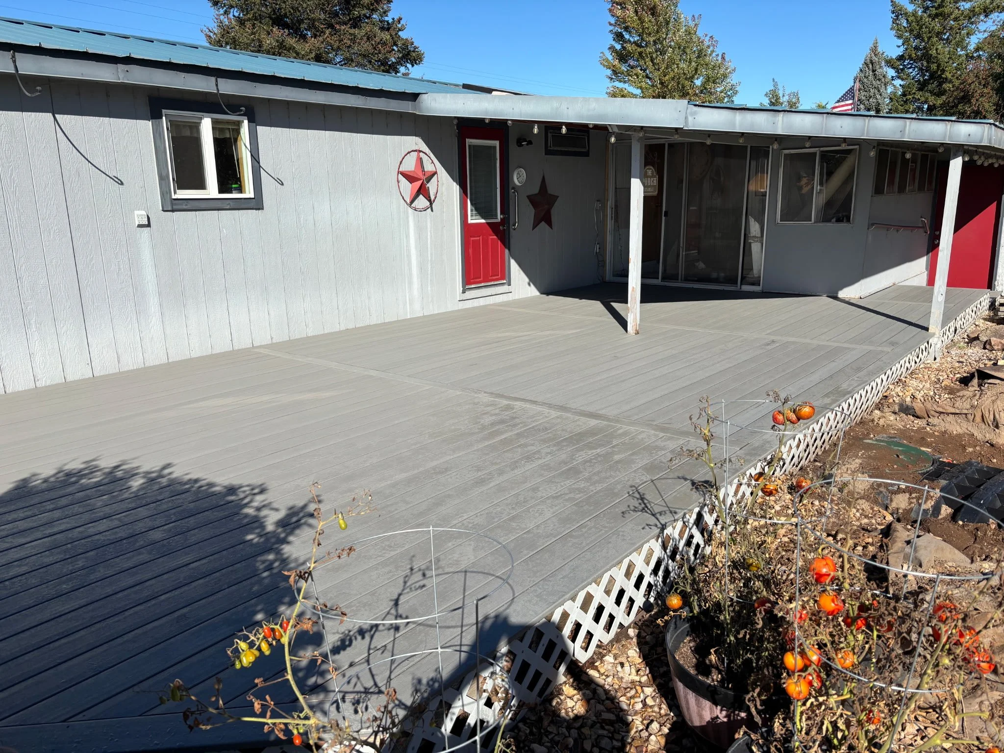 Backyard Trex deck with plants, white lattice border, gray house with red doors, and hardwired outdoor lighting fixtures, under clear blue sky and trees with autumn foliage. 