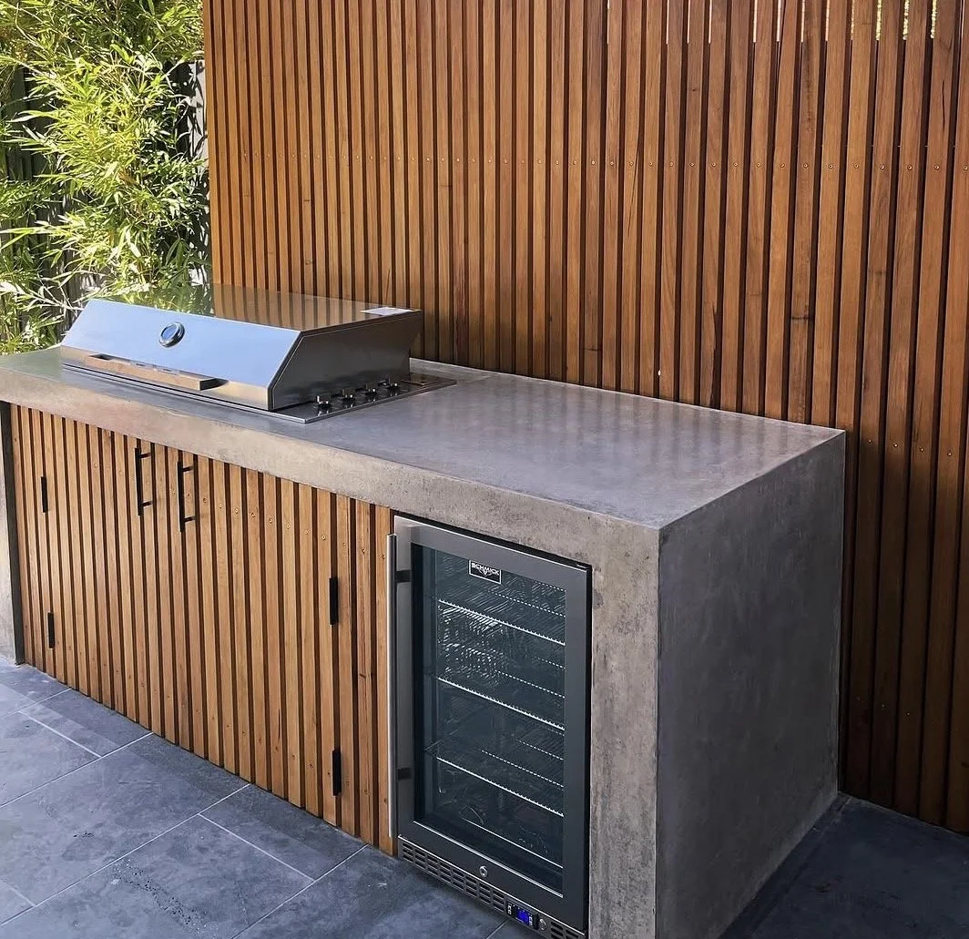 Outdoor kitchen area with a built-in grill on a concrete counter and a small wine cooler underneath, against a wooden slat wall with some green plants on the side.