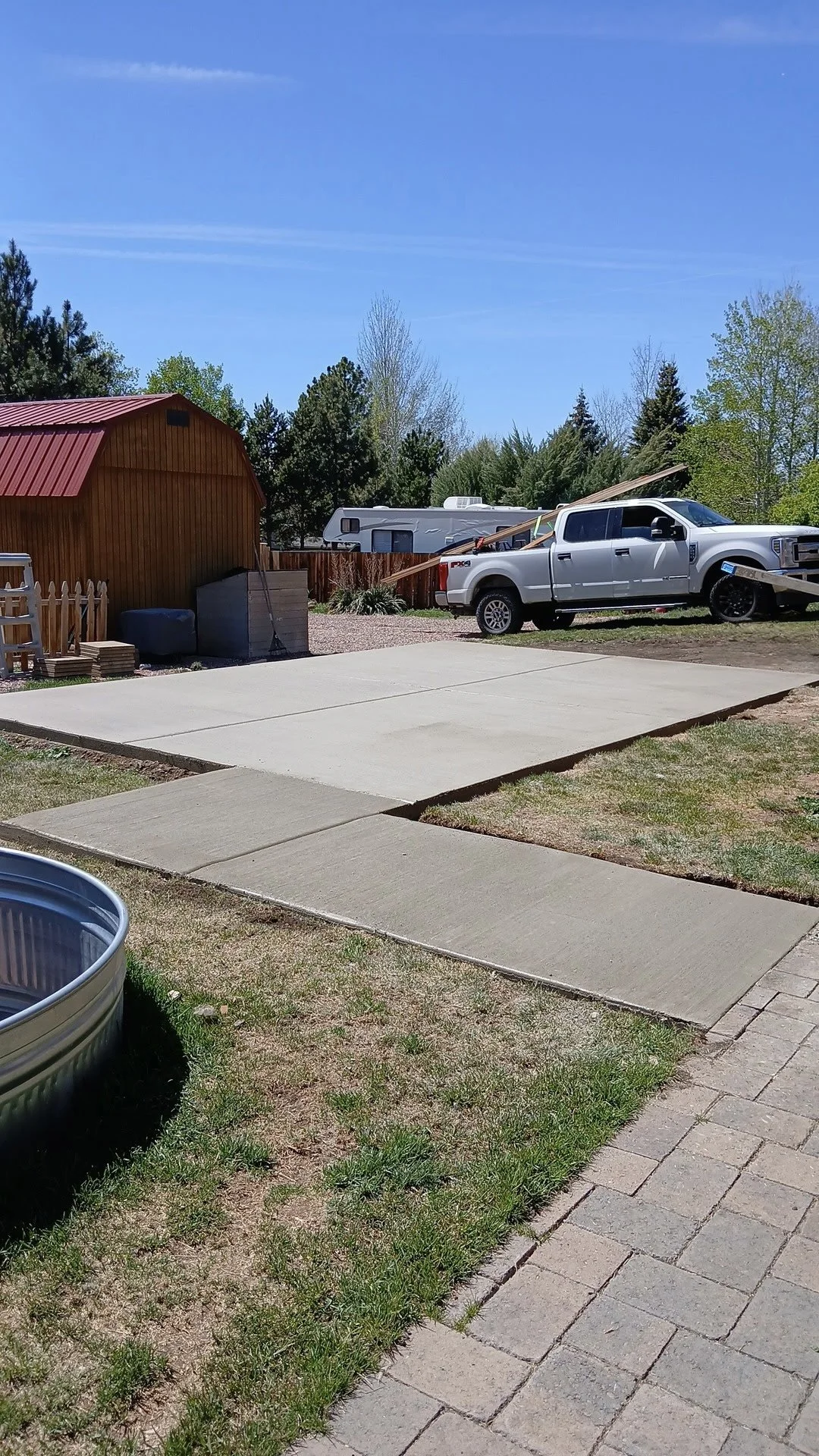 A backyard with a newly poured concrete patio, a wooden shed, a pickup truck, and a RV under a clear blue sky.