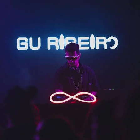 DJ wearing headphones and sunglasses performing on stage at night, with a neon infinity symbol in front and illuminated logo or text behind.
