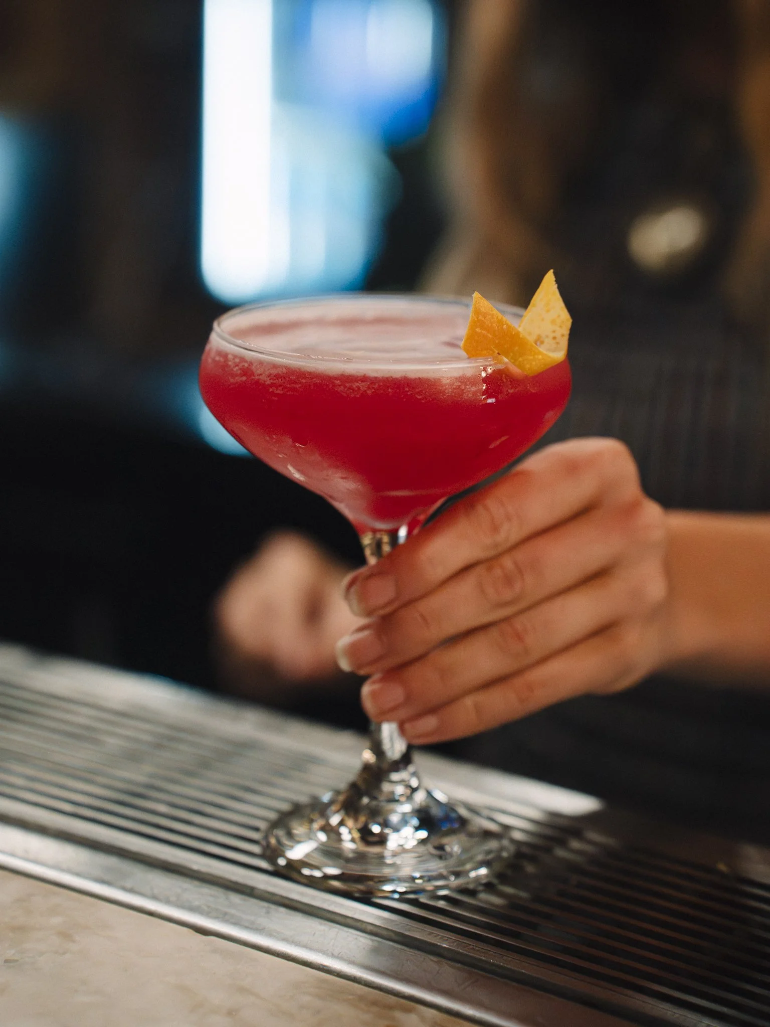 Close-up of a person's hand holding a cocktail glass with a red drink garnished with an orange peel, placed on a bar counter with a striped cloth underneath.
