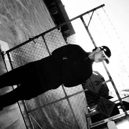 A person standing outdoors near a chain-link fence, holding a baseball bat, wearing sunglasses and a black jacket.