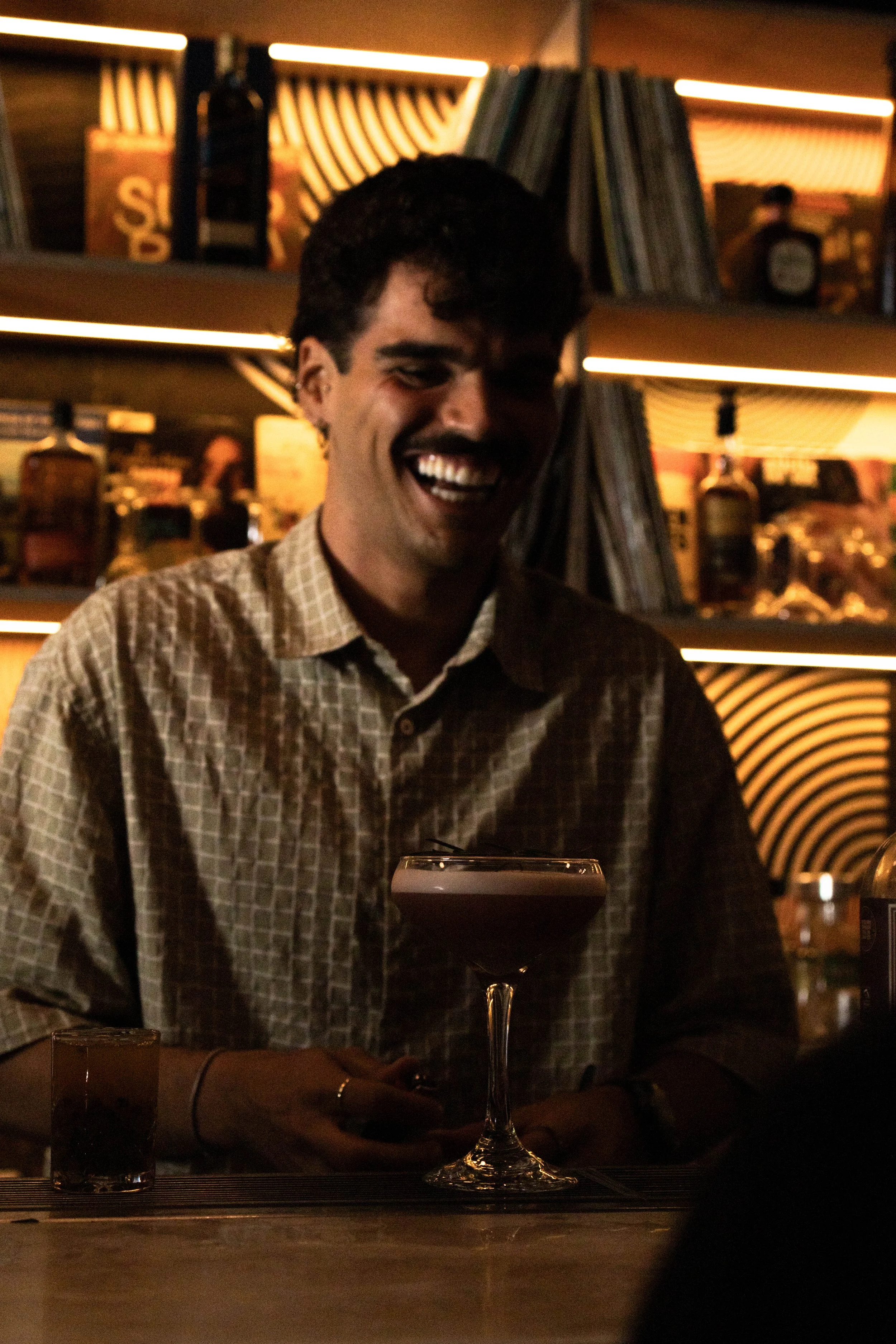 A man with dark curly hair and a mustache, smiling and looking at his phone, sits at a table in a dimly lit bar or restaurant. On the table, there is a glass with a dark drink and another glass in front of him. The background features bookshelves with books and warm lighting.