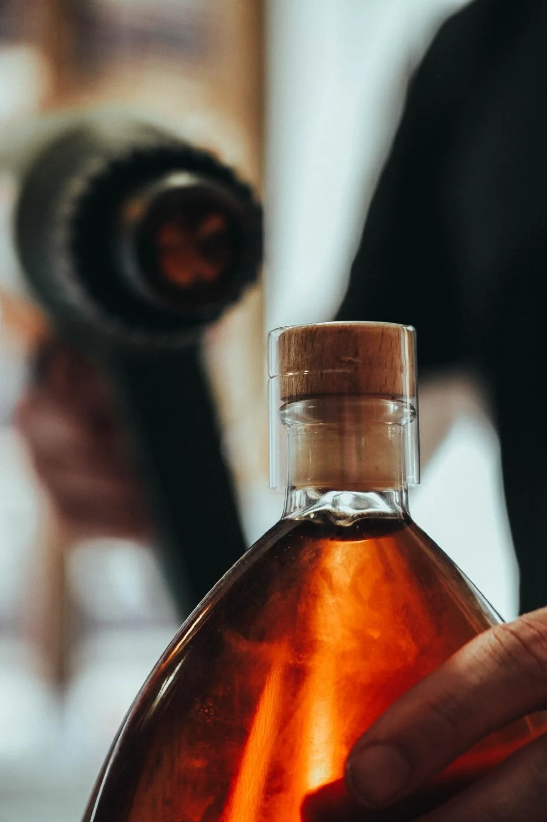 Une main tient une bouteille de spiritueux en verre brun avec un bouchon en bois, devant un fond flou où une personne regarde par un microscope.
