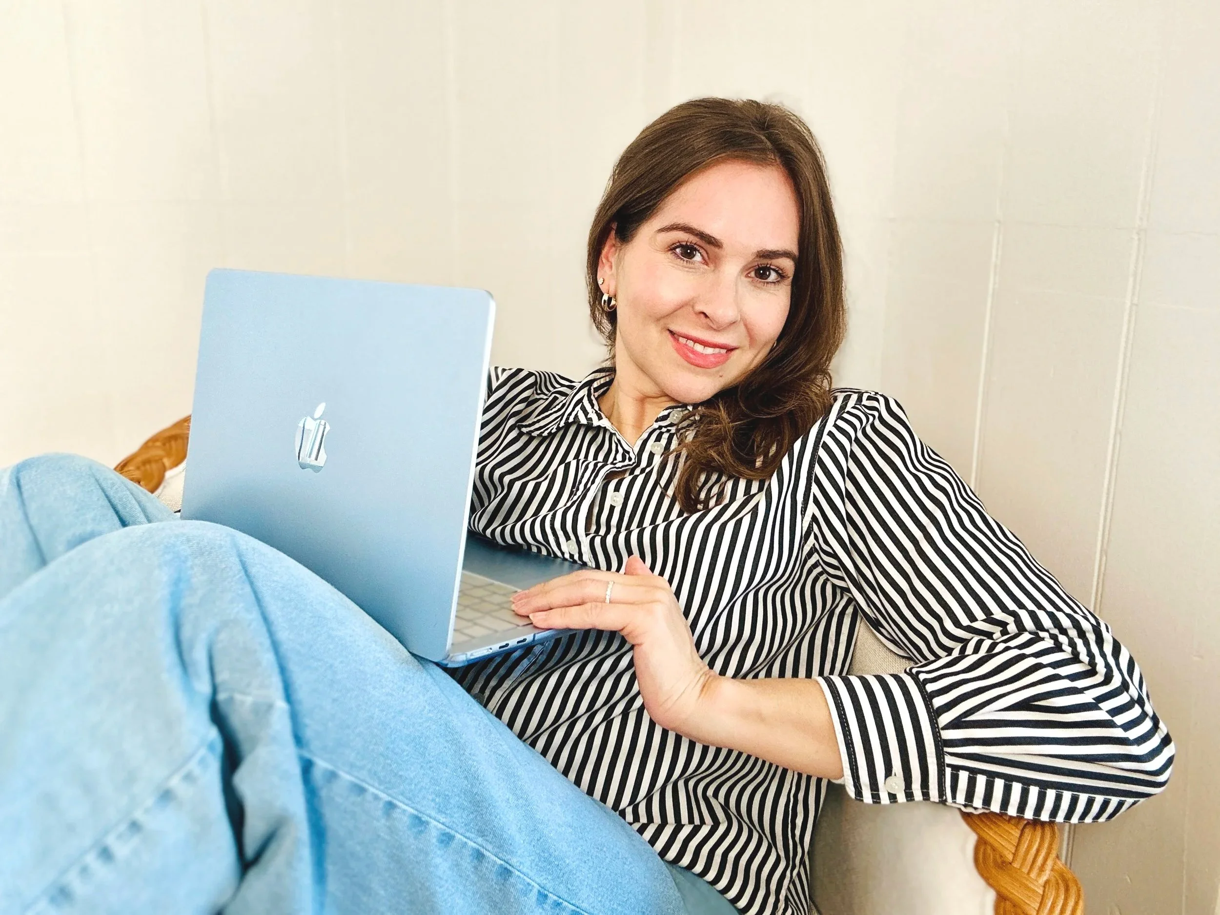 A woman with brown hair, wearing a black and white striped shirt, smiles while sitting on a beige chair with a wooden armrest, next to a light blue MacBook, in front of a plain light-colored wall.