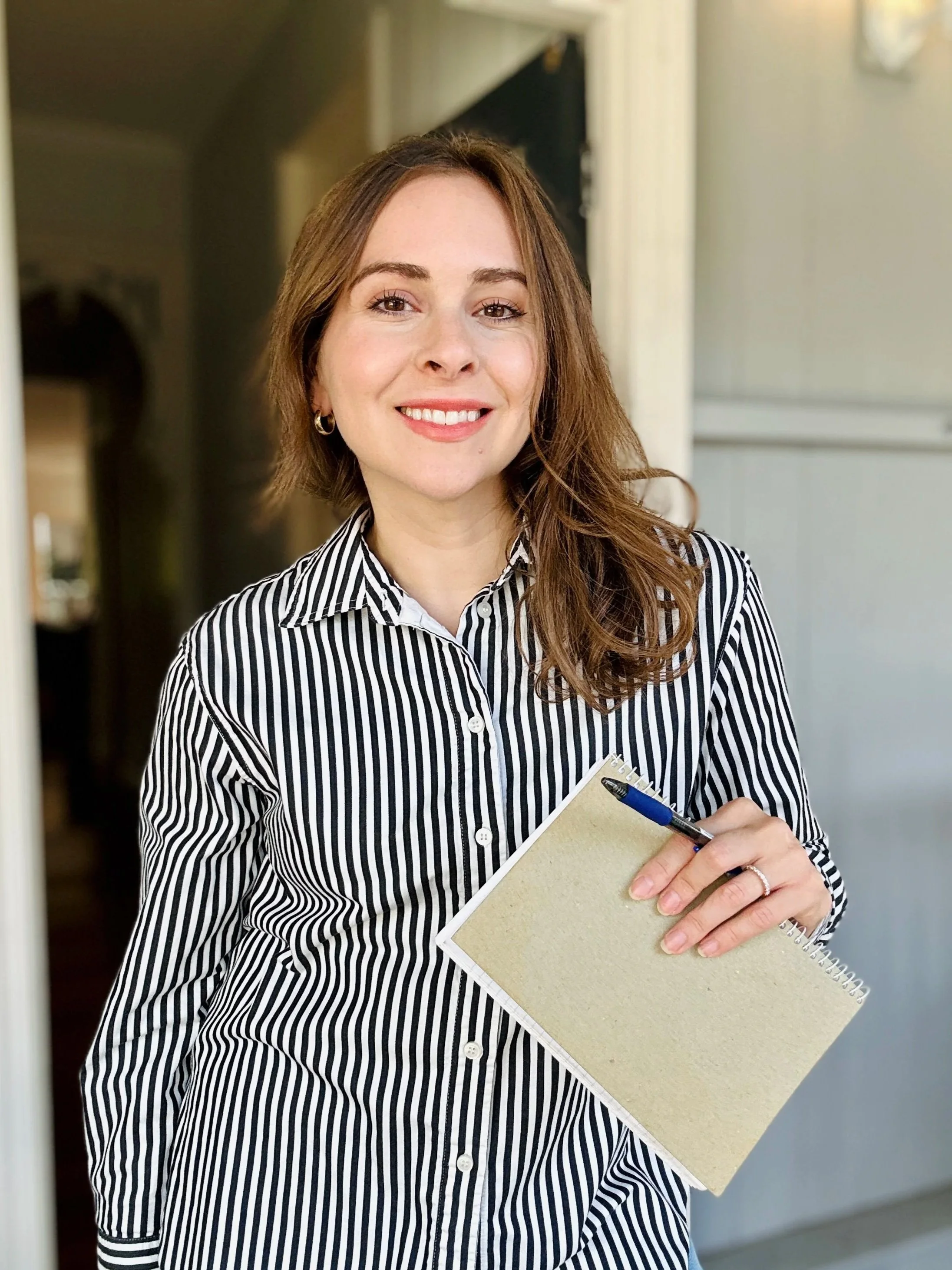 A woman with brown hair, wearing a black and white striped shirt, holding a small notebook and pen, smiling in an indoor setting.