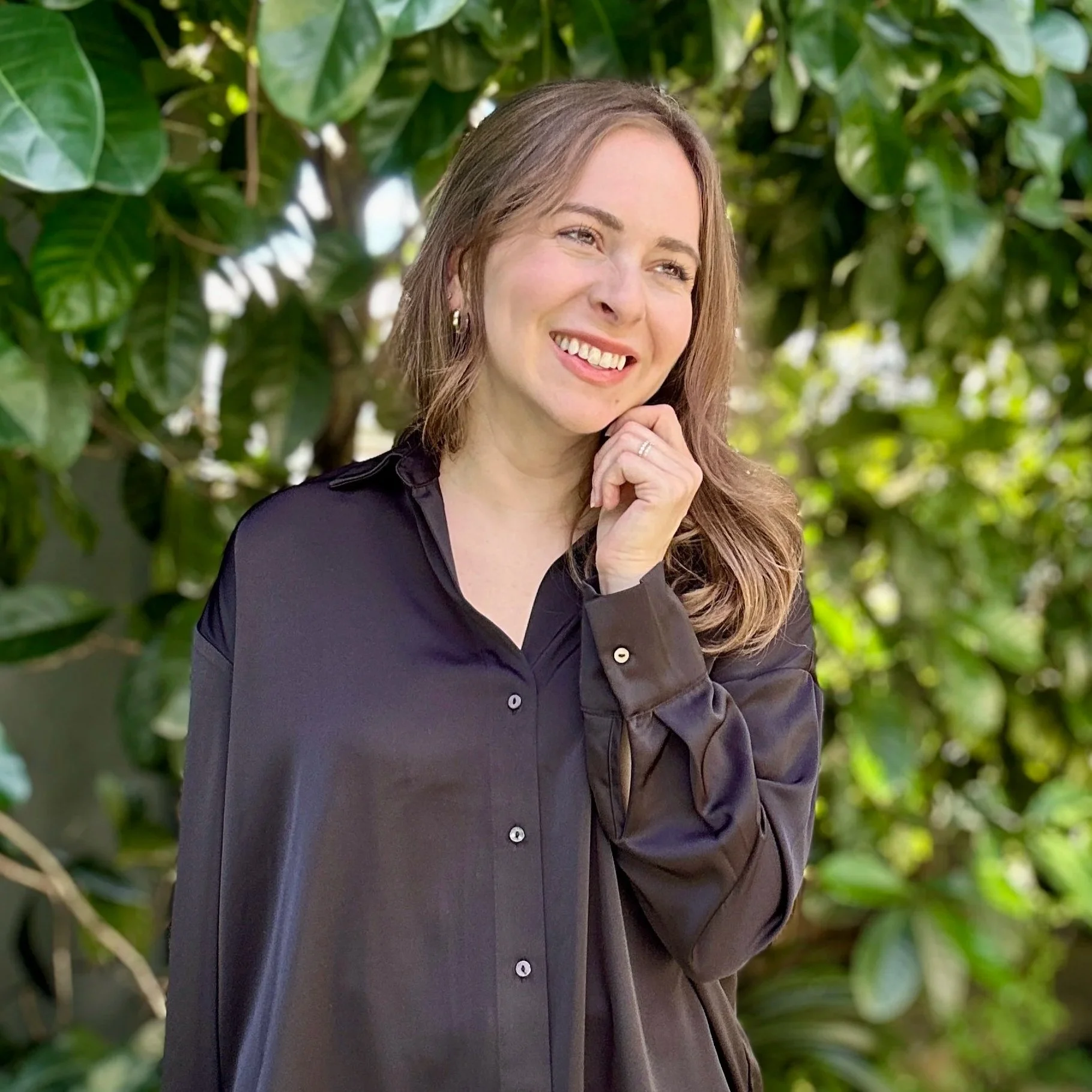 A woman with long, wavy brown hair, smiling and touching her chin, stands outdoors in front of green leafy foliage, wearing a black shirt.
