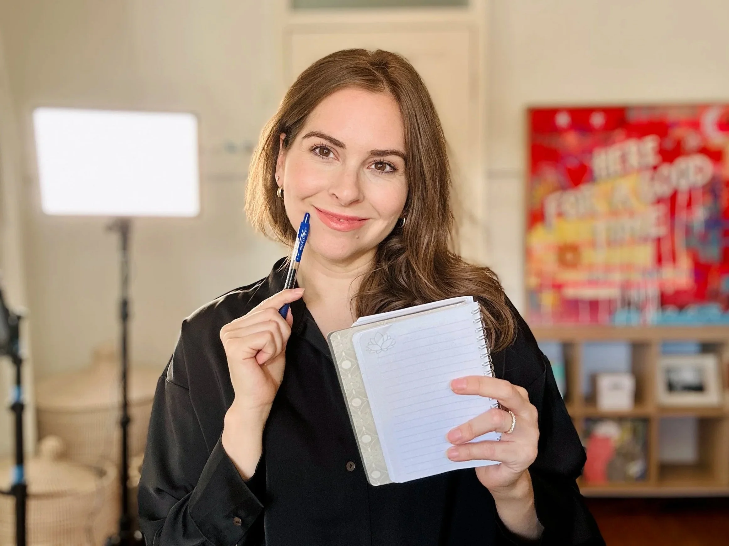 A woman with long brown hair and earrings is smiling, holding a blue pen to her cheek, and a notebook in front of her, standing in what appears to be a library or classroom with books in the background.