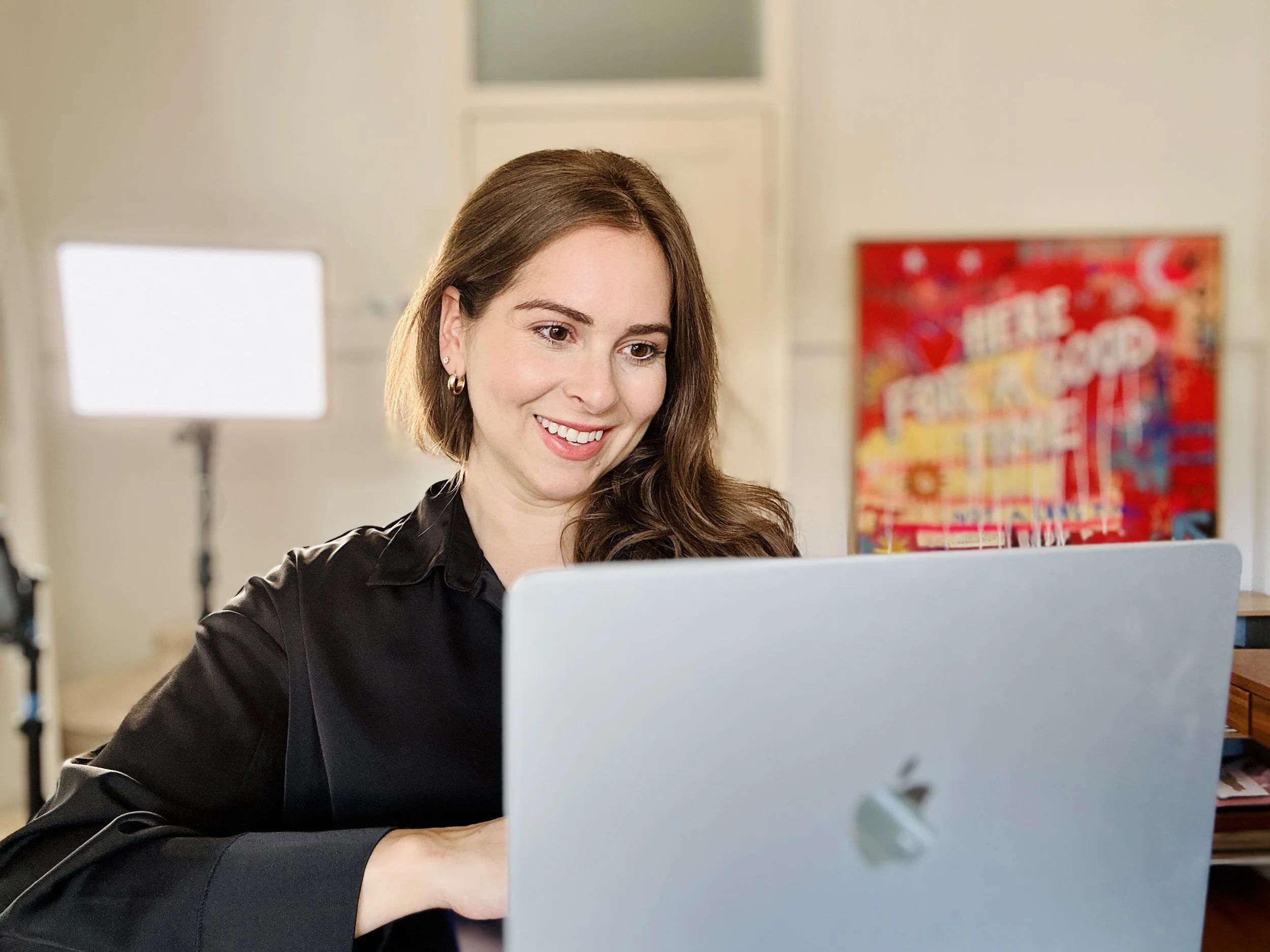A woman with long brown hair and earrings smiling while working on a laptop in a brightly lit room with a colorful poster in the background.