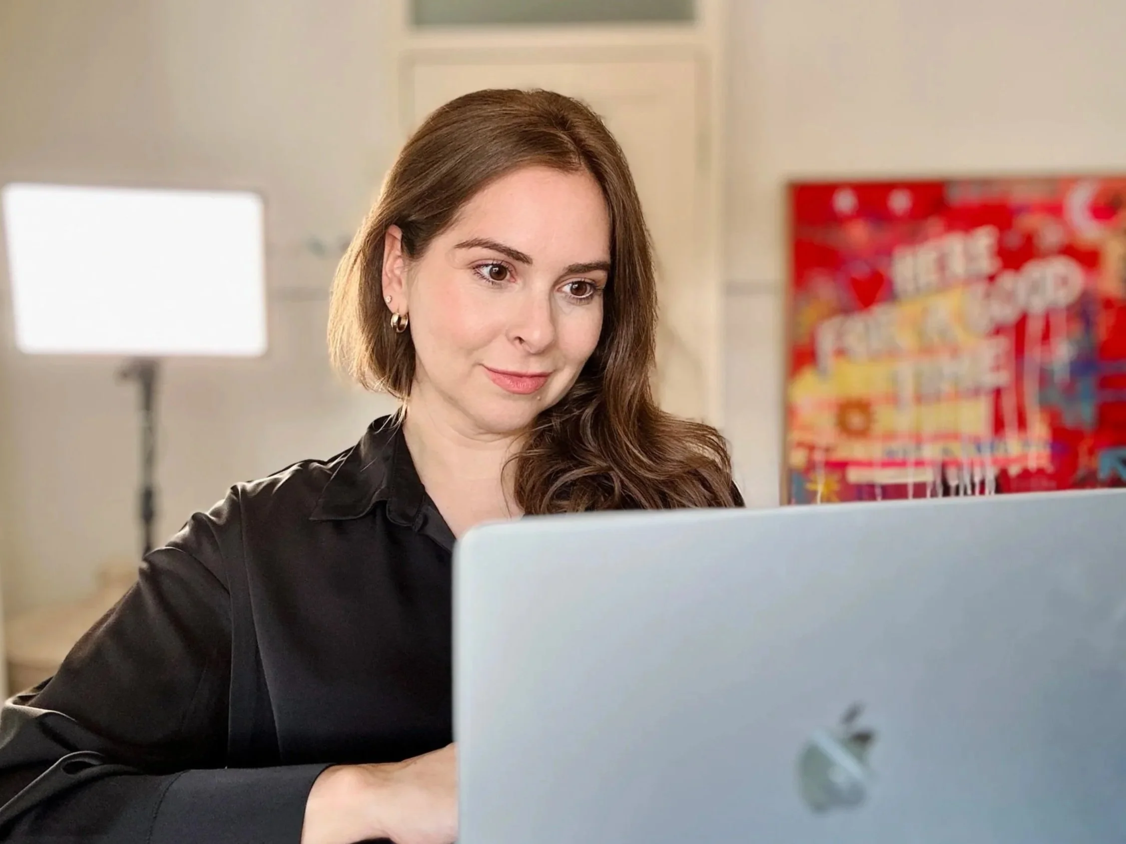 A young woman with brown hair and hoop earrings looks at a laptop screen in a well-lit room with a blurred colorful poster in the background.