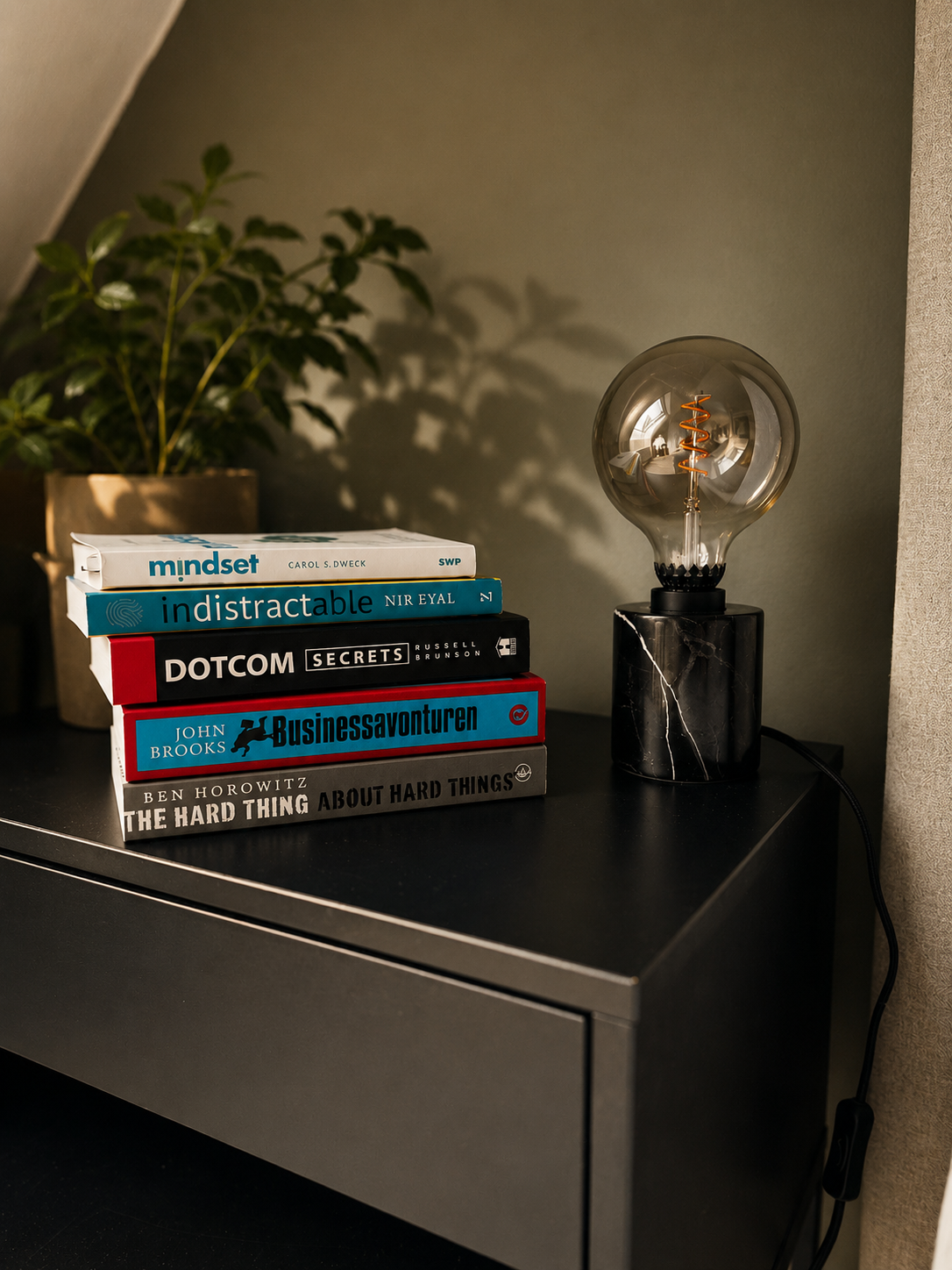 Stapel boeken op een zwart bureau naast een moderne lamp met glazen bol en een zwarte marmeren voet, met een groene plant op de achtergrond.