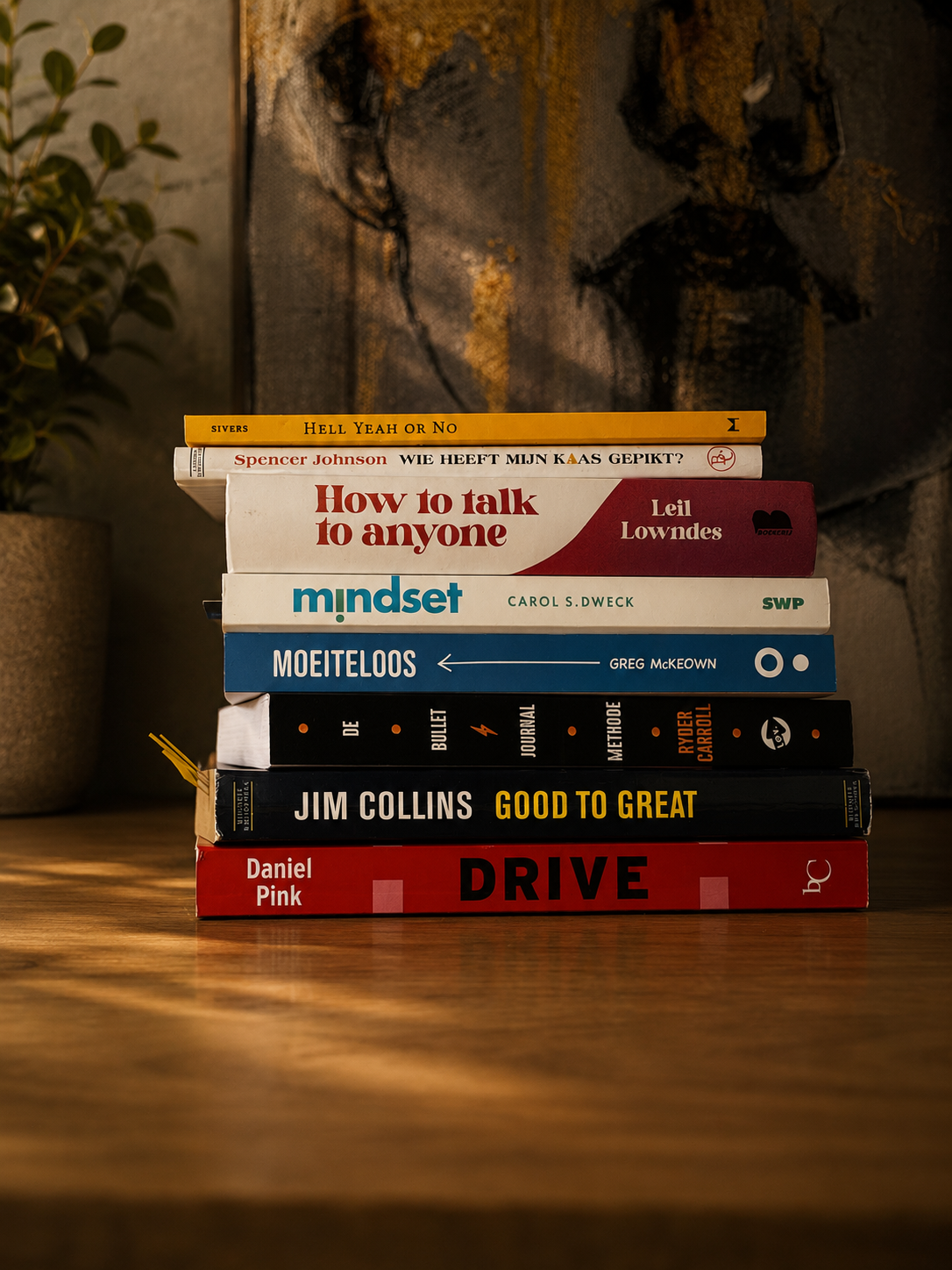 Een stapel boeken op een houten tafel, met een plantenpot aan de linkerkant en een kunstwerk op de muur op de achtergrond.