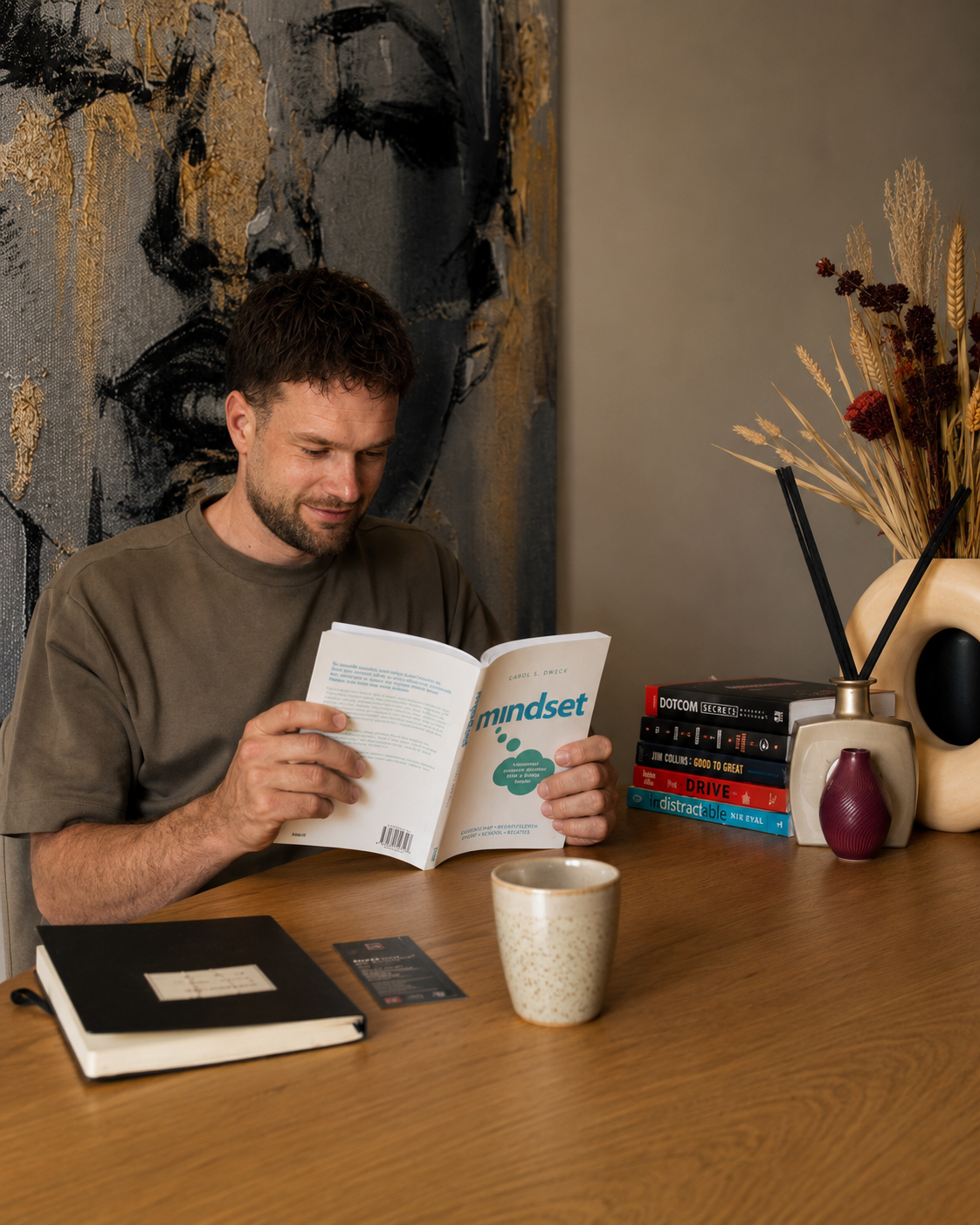 Man zit aan een houten tafel en leest een boek met de titel 'mindset'. Op de tafel liggen een zwart notitieboek en een bankpas, terwijl er een beker en een vaas met gedroogde bloemen op de tafel staan. Op de achtergrond hangt een abstract kunstwerk.