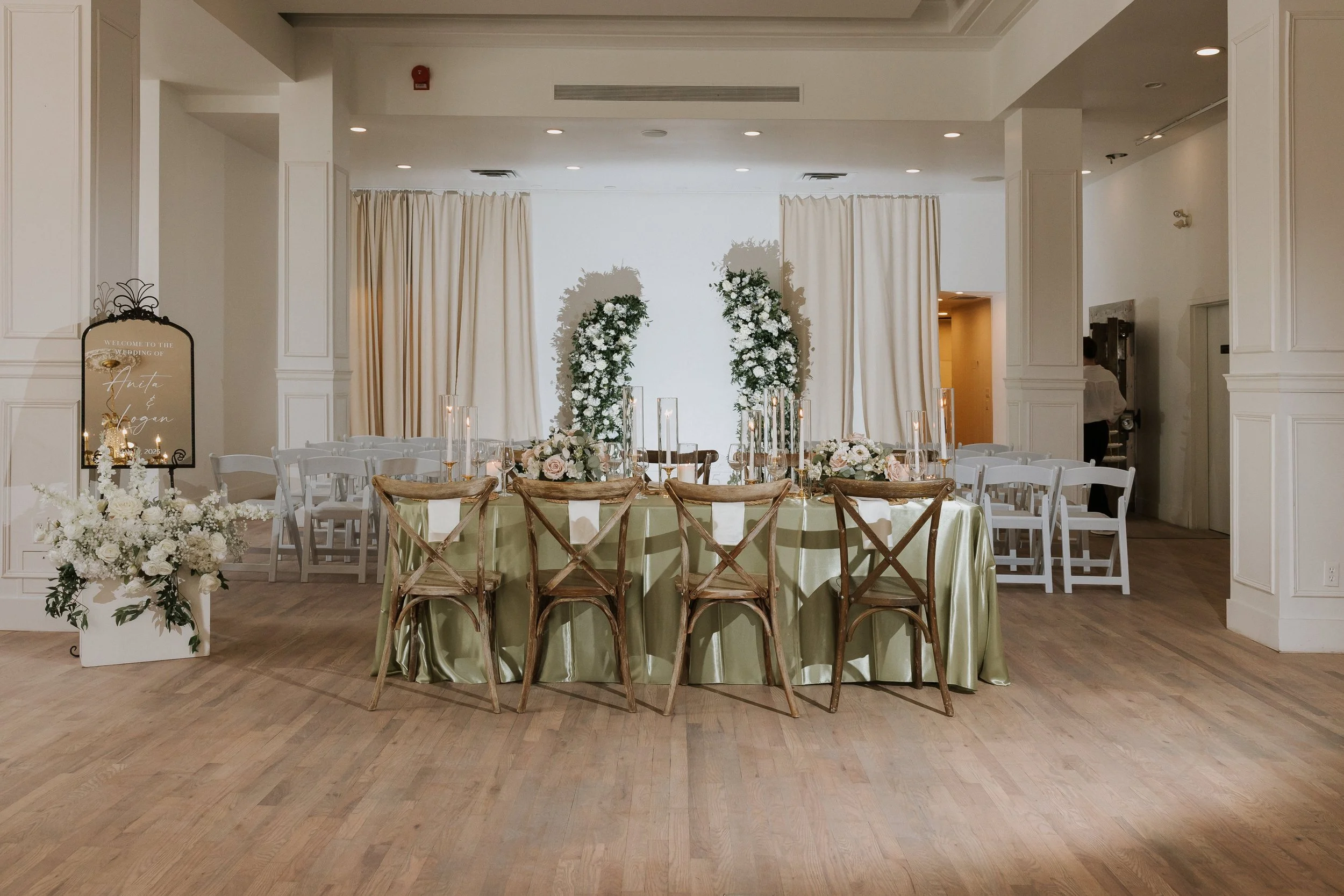 Wedding reception setup with a head table decorated with white flowers and tall candles, white chairs, large floral arrangements, and a welcome sign for the wedding of Anita and Hagen.
