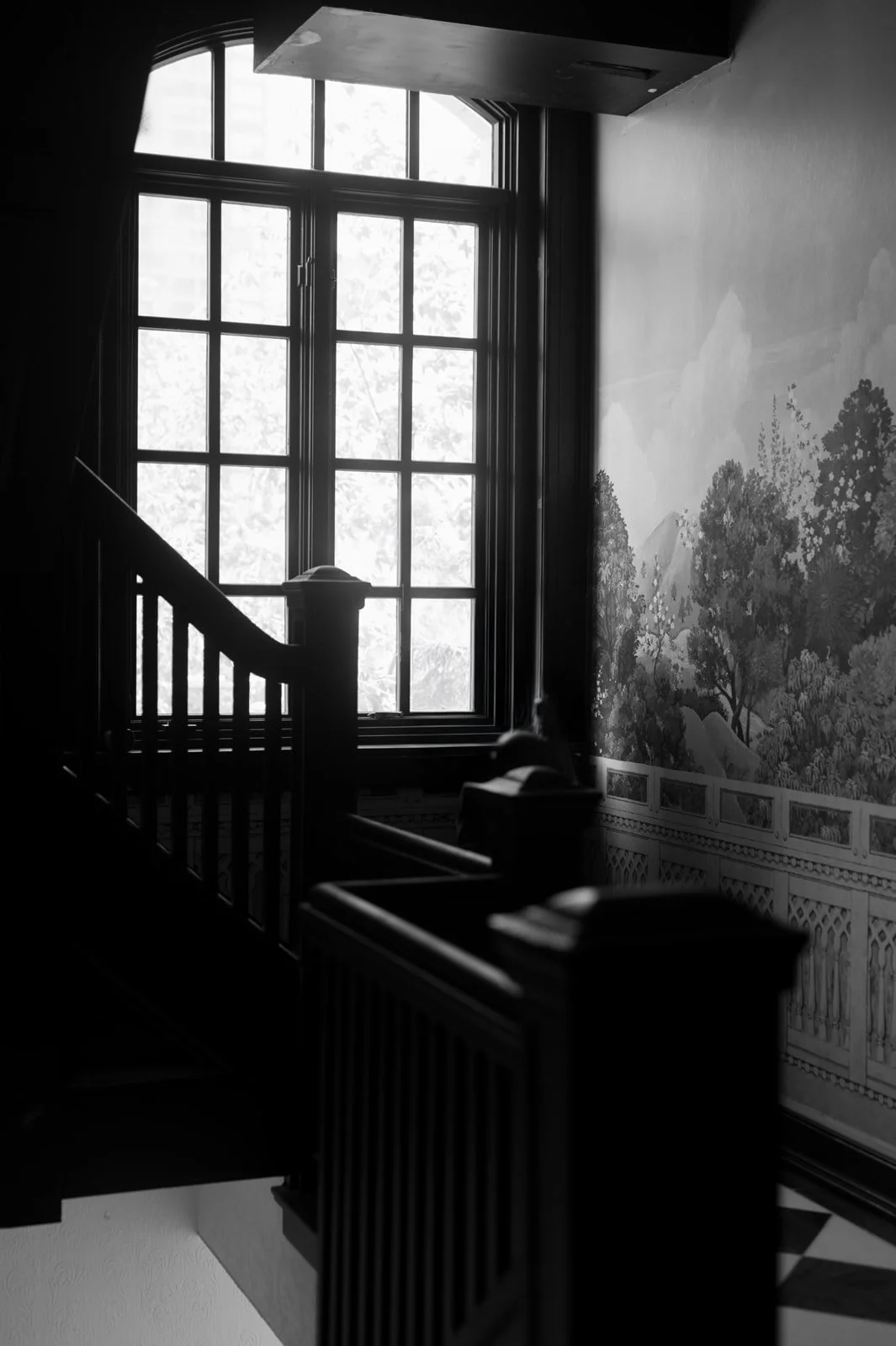 Black and white photo of a staircase and a large window, revealing a painted landscape mural on the wall.