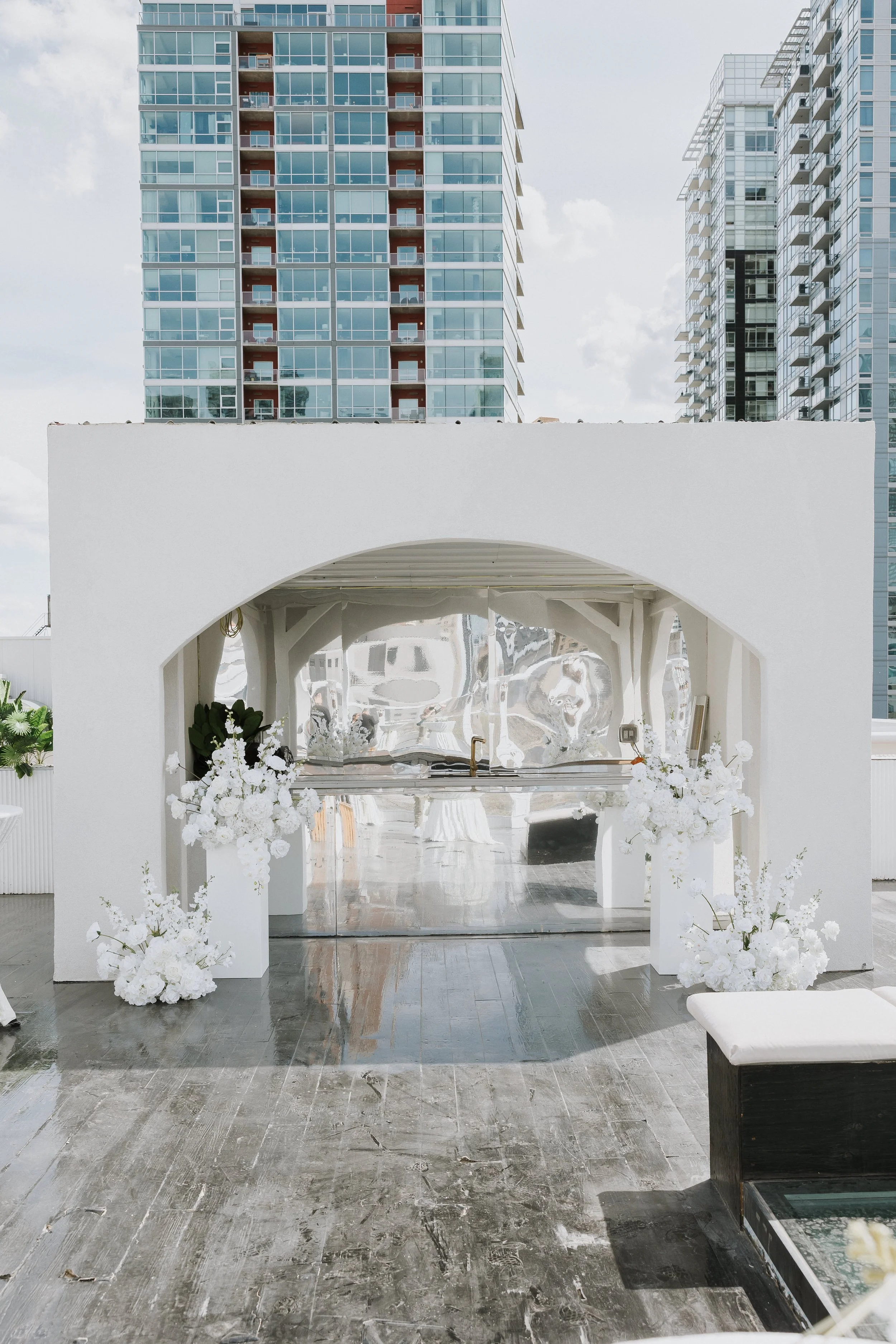 Modern outdoor event space with a white archway and floral arrangements, city skyscrapers in the background, and a reflective gray stone floor.