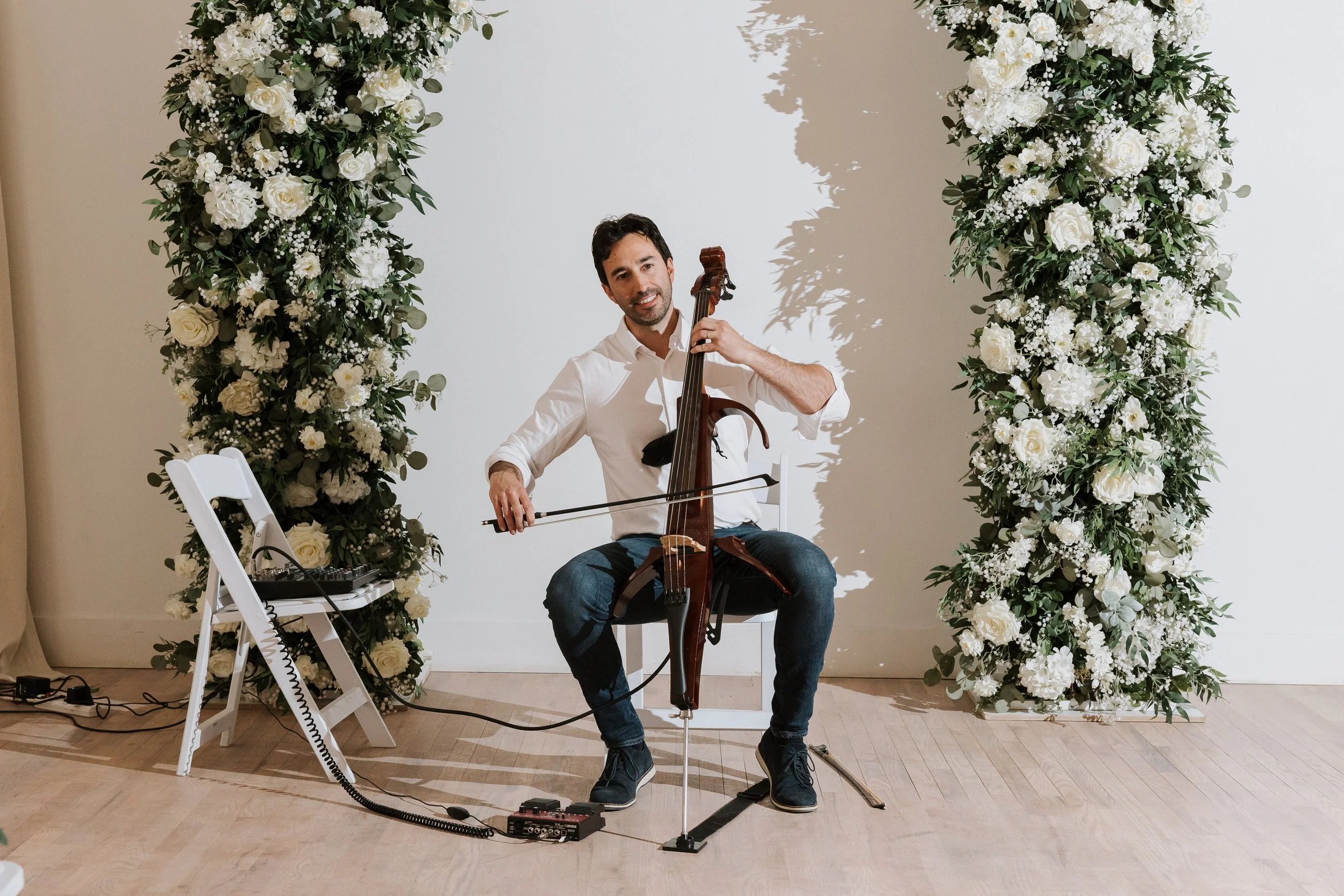 A man playing a cello at a wedding or event with white floral arch behind him