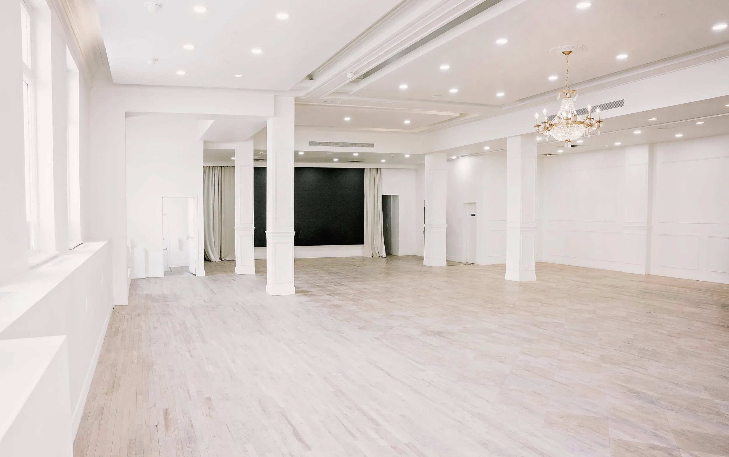 An empty event hall with white walls, light wood flooring, a chandelier, several ceiling lights, tall windows, and a black accent wall with white curtains.