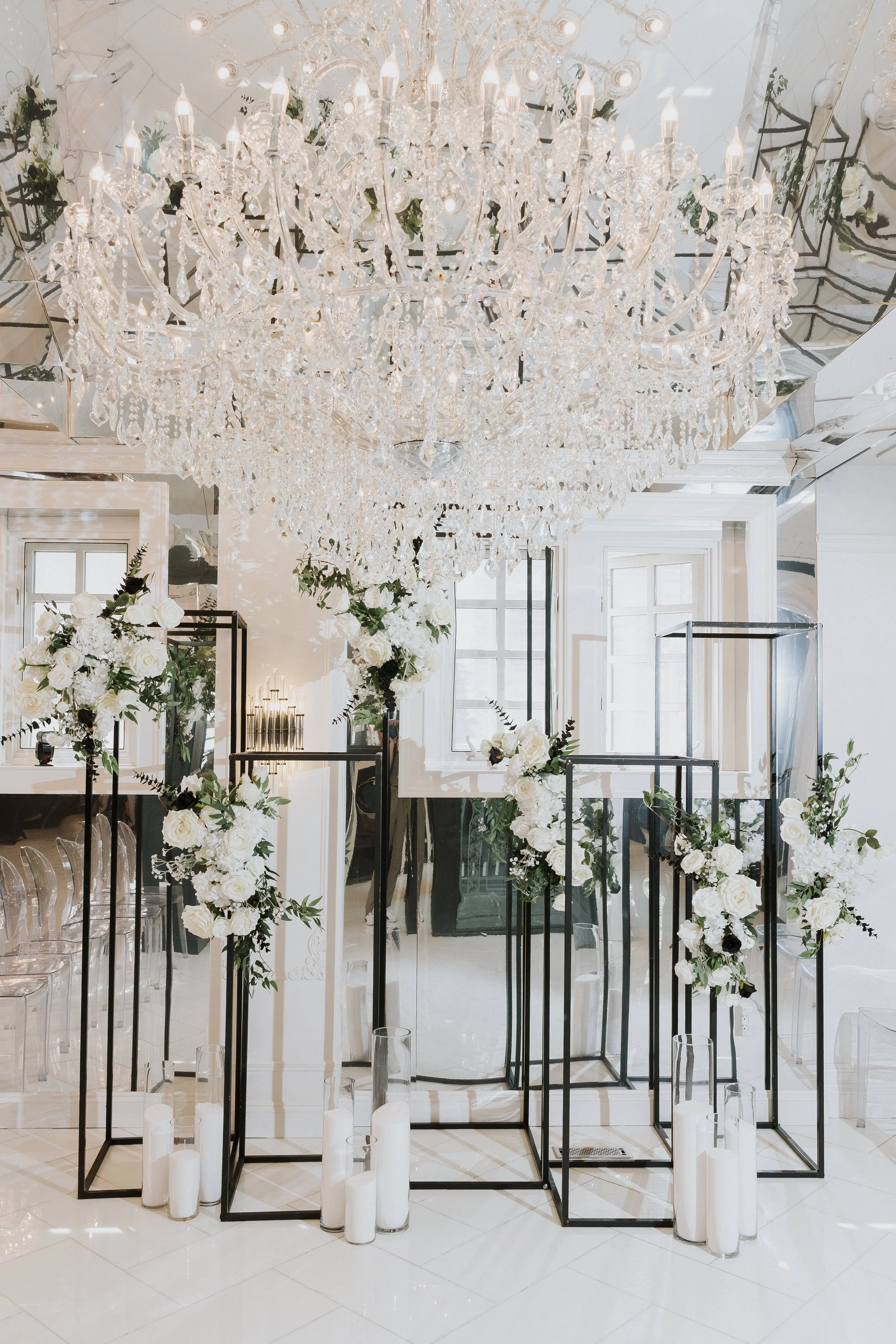 Elegant interior with a large crystal chandelier, black metal display stands with white floral arrangements, and tall white candles in glass holders on a white tiled floor.