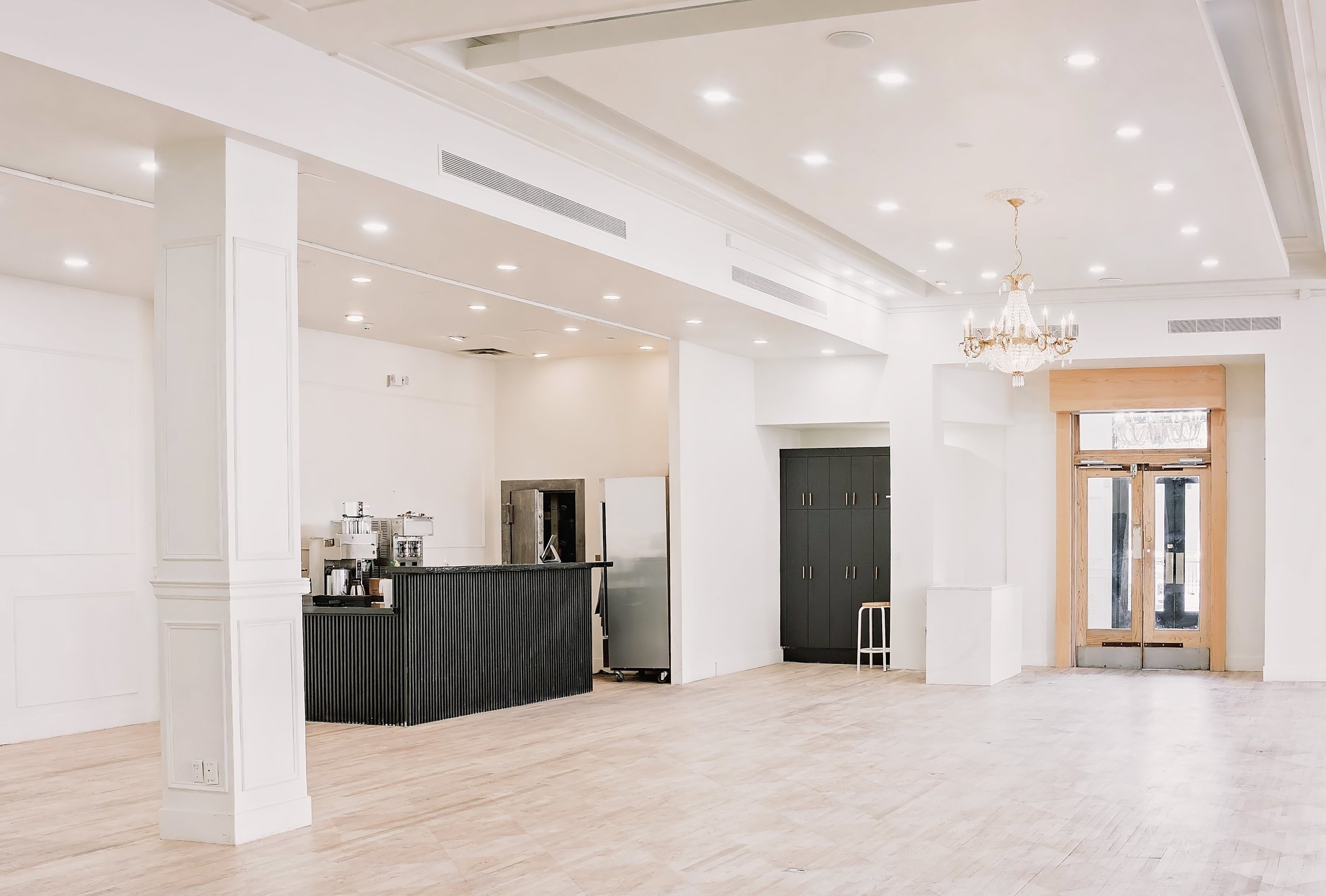 Empty interior space with white walls, light wood flooring, a black curved reception counter, and a chandelier hanging from the ceiling near a large wooden-framed glass door.