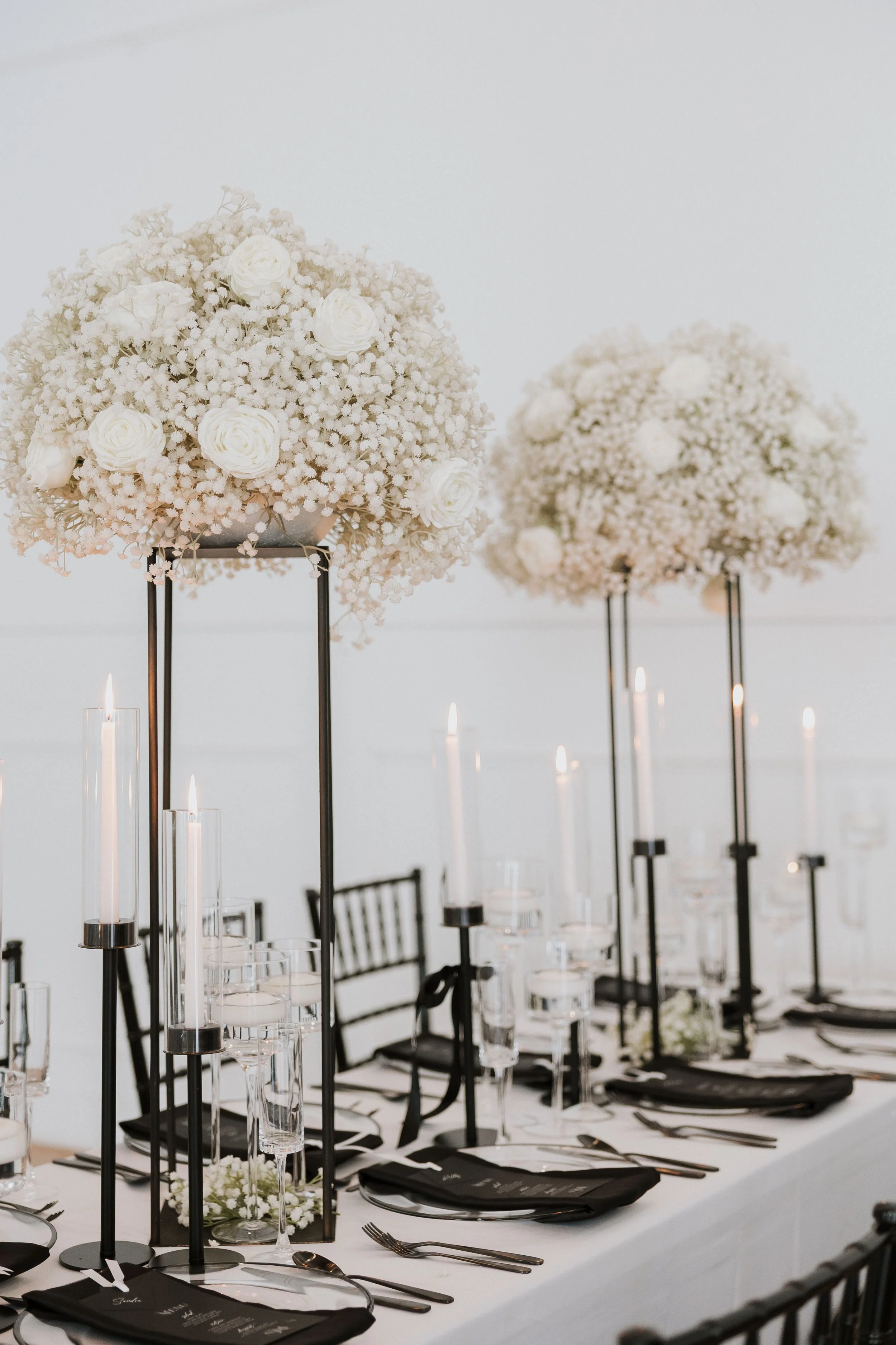 Elegant wedding reception table setup with tall floral centerpieces of white roses and baby's breath, black chairs, and candles.