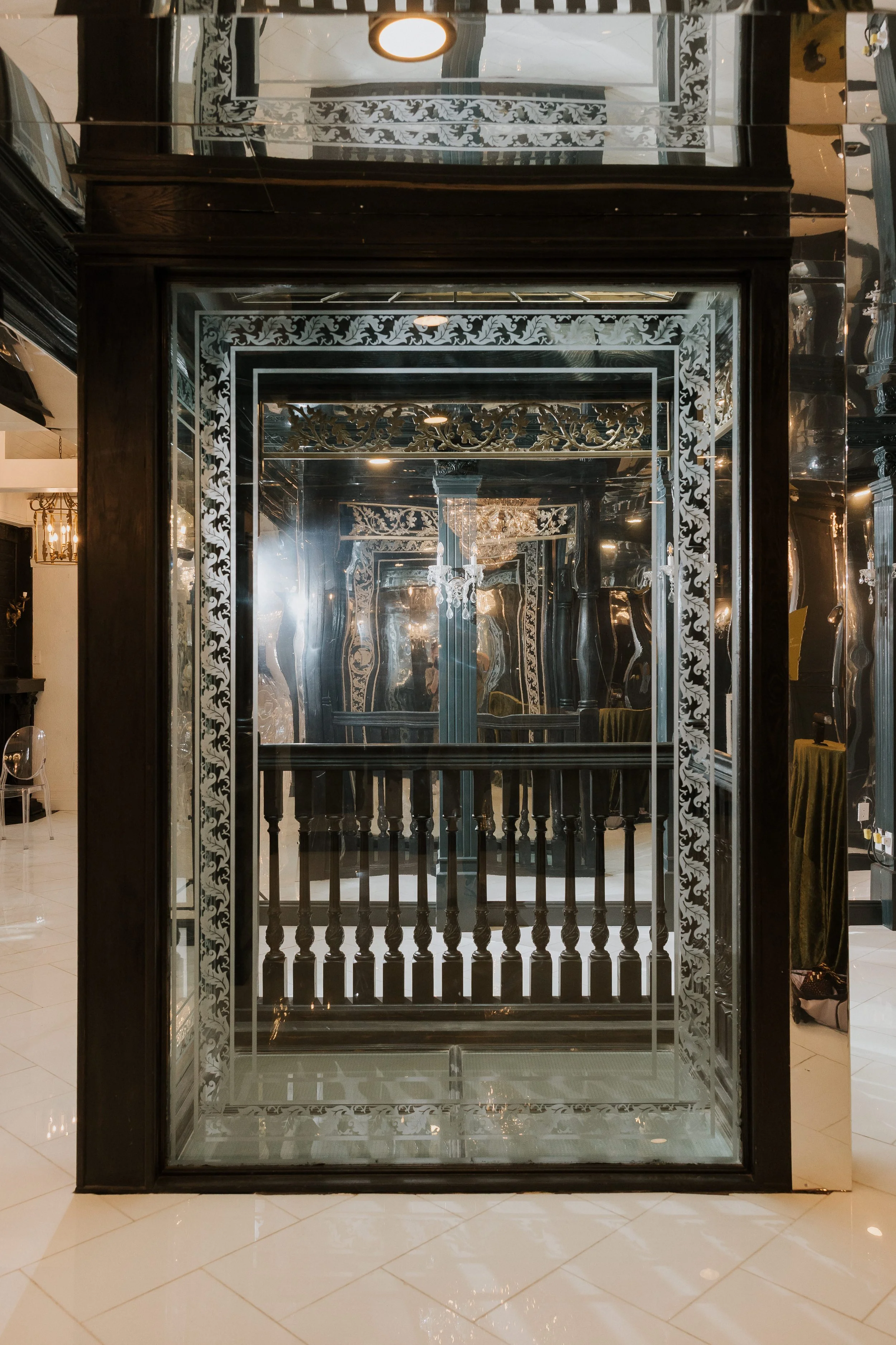 A mirrored elevator door with black framed ornamented patterns. The reflection shows a chandelier and decorative ceiling details inside the elevator.