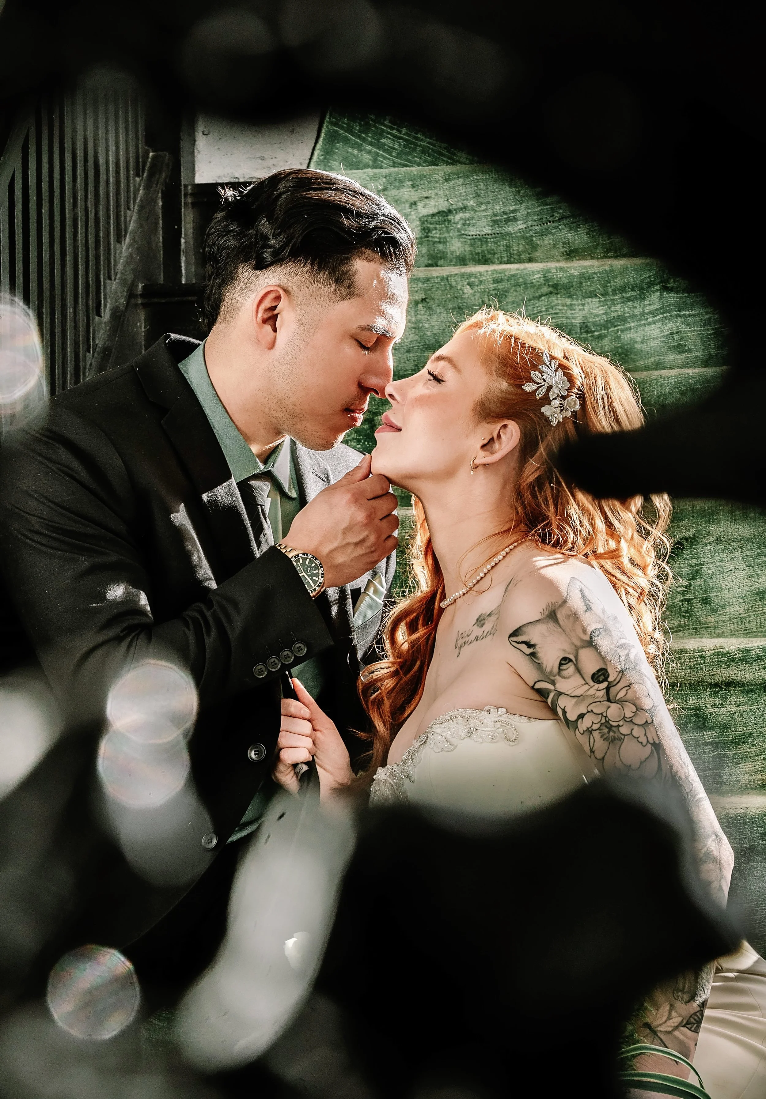 A bride and groom holding hands, leaning close with their noses touching, in a romantic moment during their wedding. The groom is in a black suit, and the bride has red hair with a decorative hairpiece and visible tattoos on her shoulder. The photogr