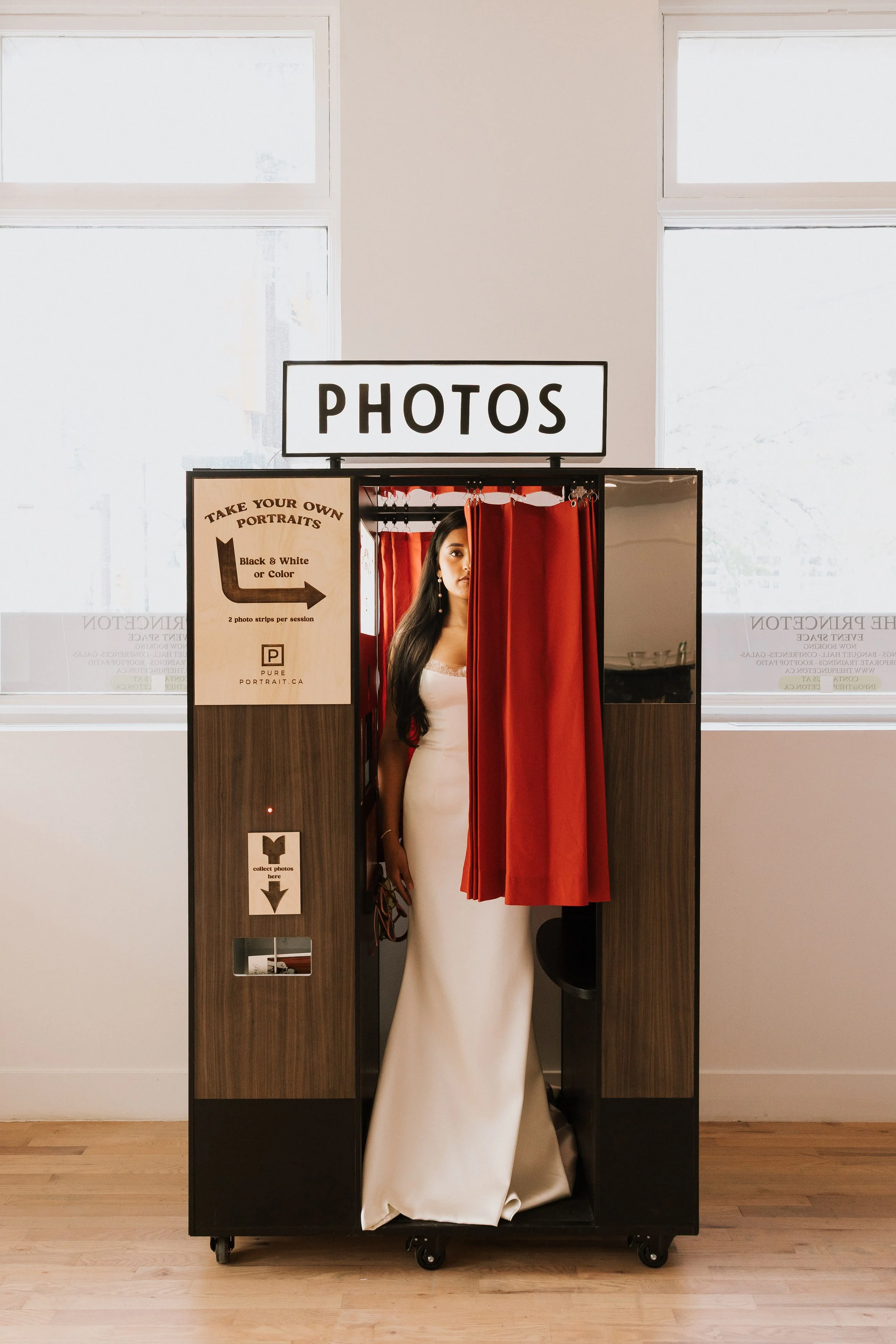 Photo booth with a sign that says "PHOTOS" on top, featuring a person in a white dress standing behind red curtains. There are signs on the booth offering to take portraits in black and white or color, with instructions to take two photo strips per s