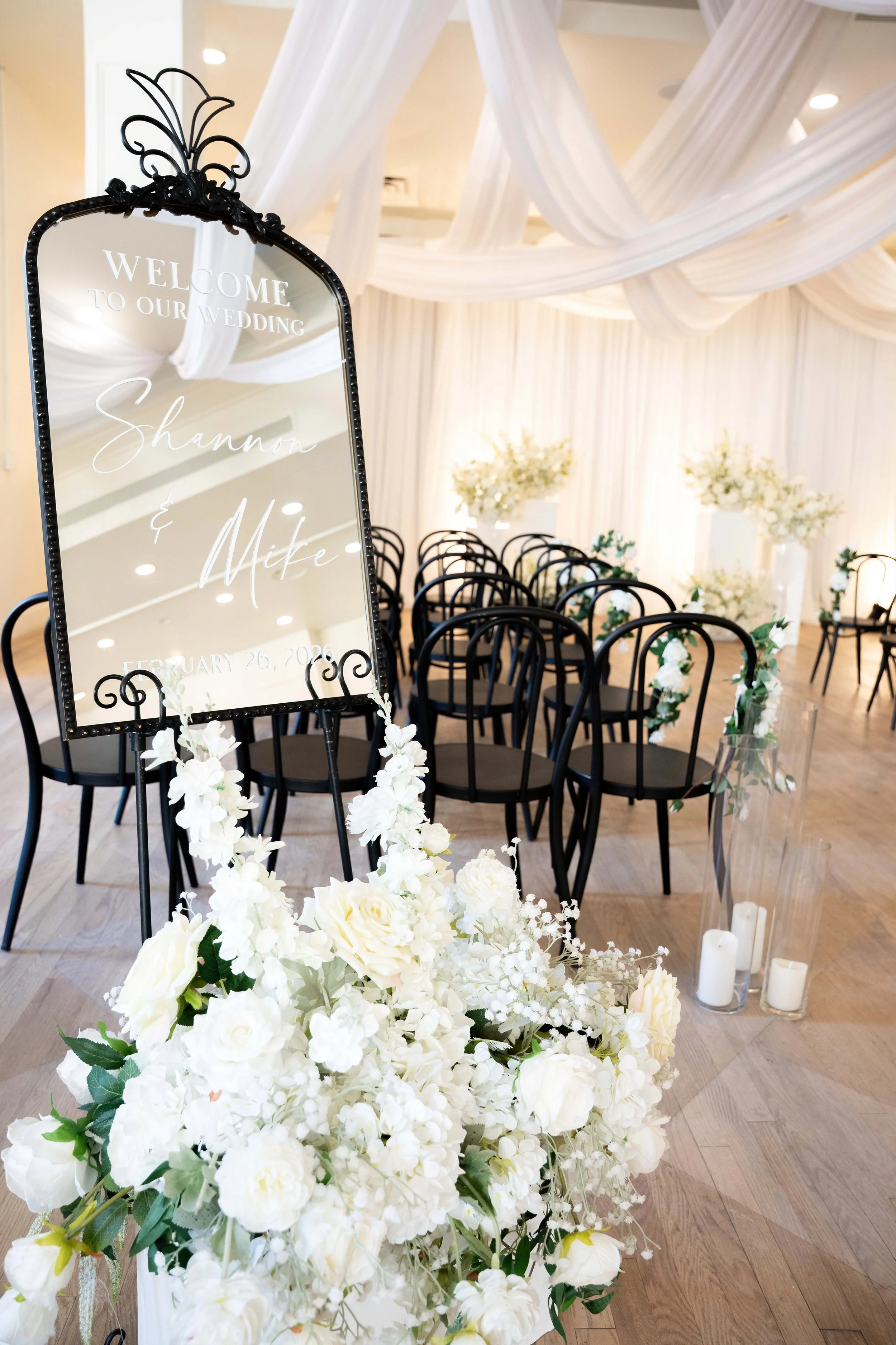 Elegant wedding ceremony setup with black chairs decorated with white flowers, a floral arrangement, and a mirror with welcoming text for Shannon and Mike's wedding on February 26, 2026, in a softly lit venue with draped white fabric ceilings.