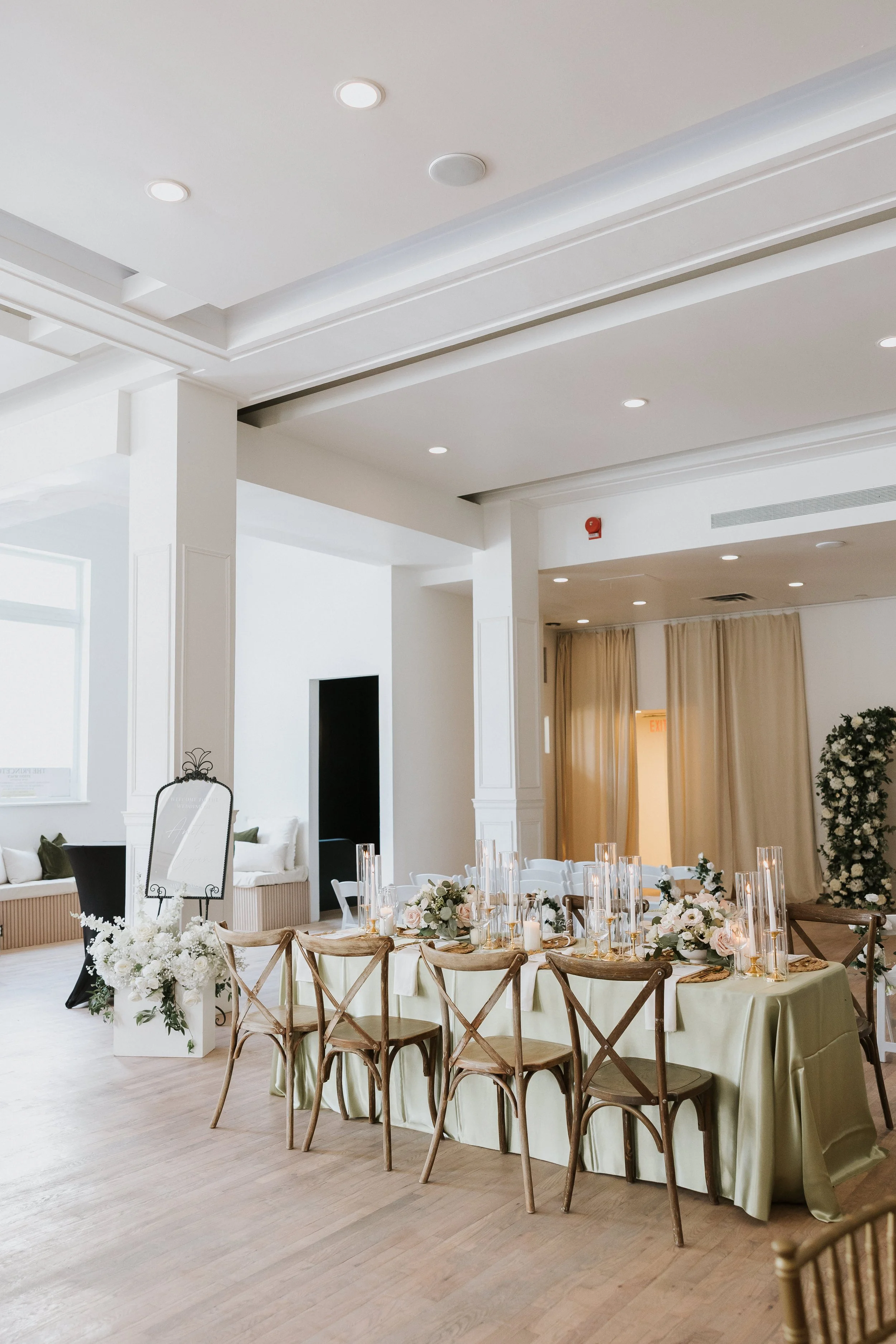 Elegant indoor event space with round dining table, chairs, floral centerpieces, and candles, featuring white walls, high ceilings, and soft lighting.