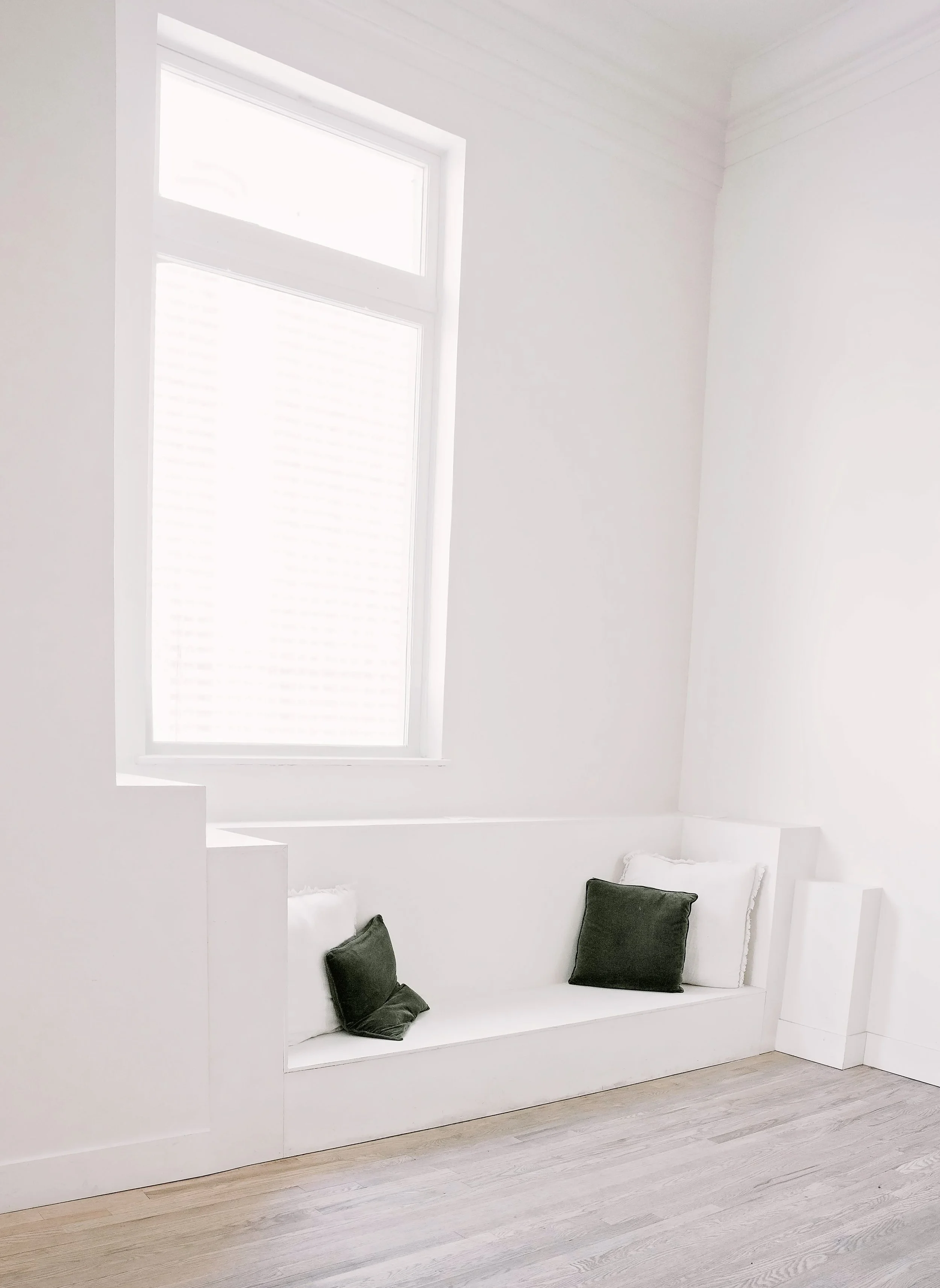 A white interior room with a large slanted window, white walls, and a seating area with four black and white pillows on a built-in cushion.