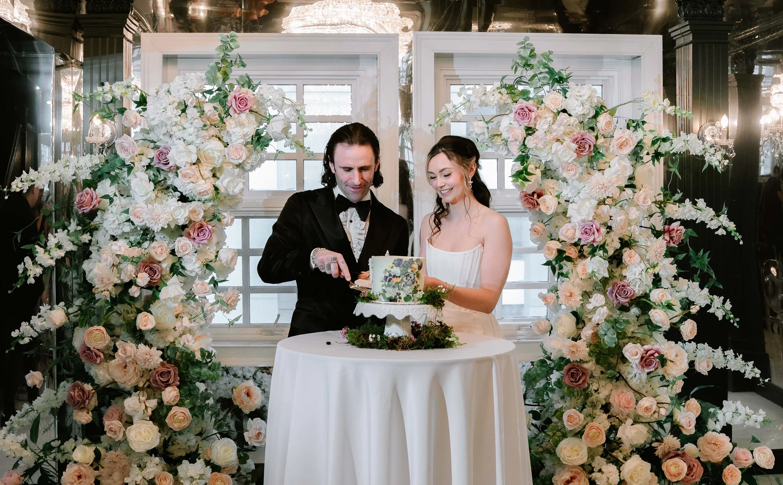 A bride and groom cutting a wedding cake, surrounded by an arch of white and pink flowers in a decorated indoor venue.