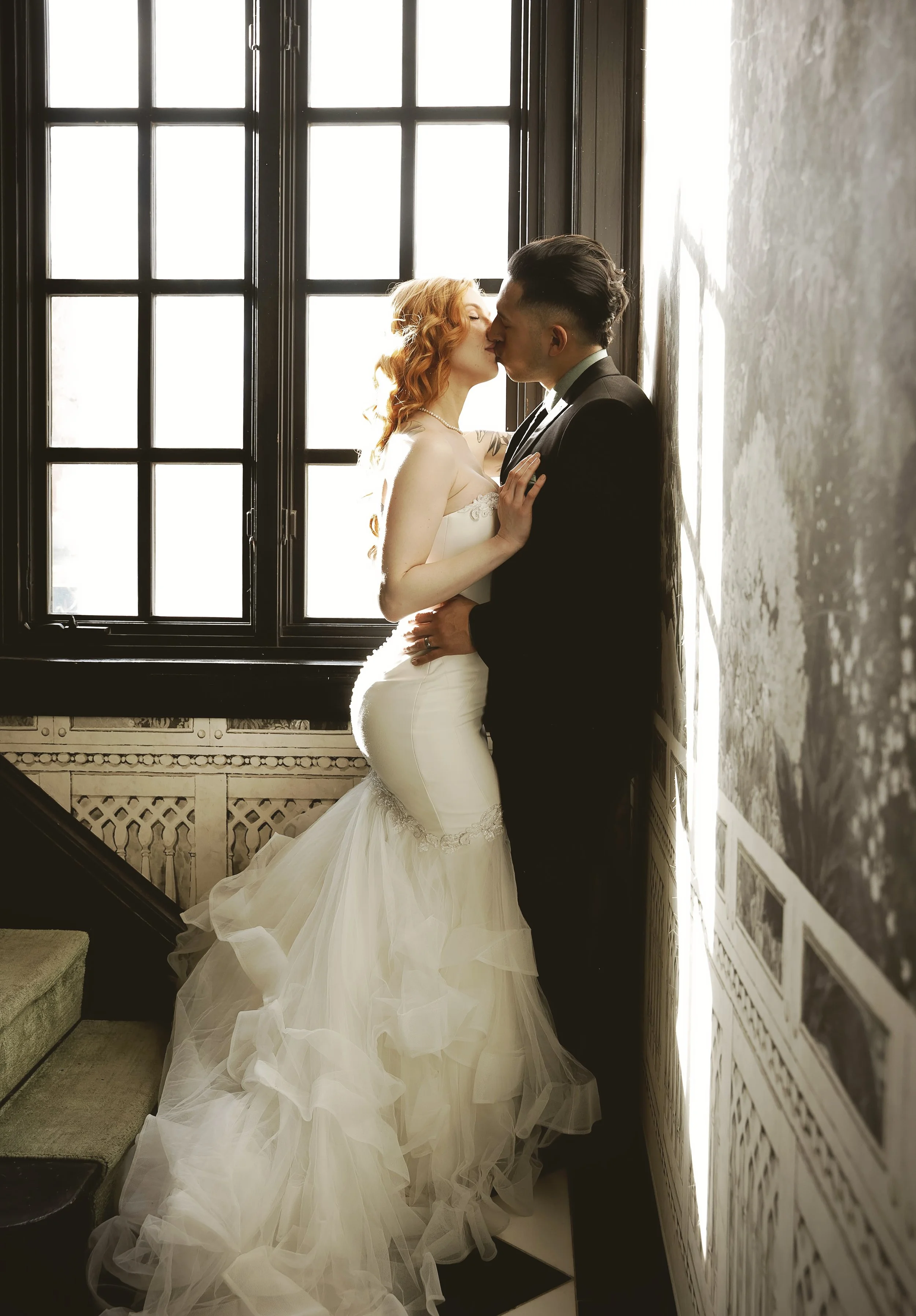 A bride and groom sharing a kiss near a window, with the bride wearing a white wedding dress with a train and the groom in a black tuxedo.