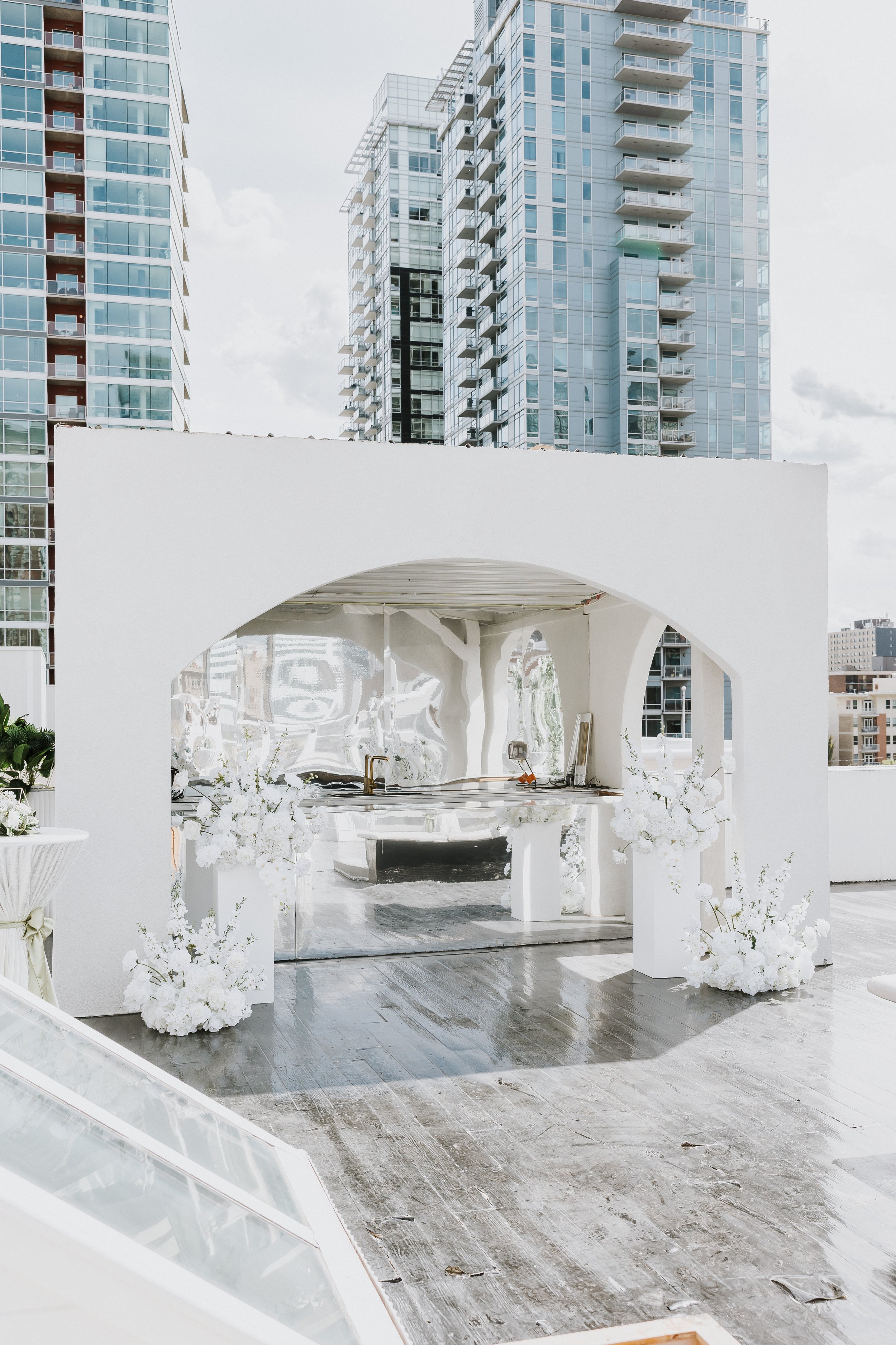 Modern outdoor wedding altar with white floral arrangements, situated on a rooftop with city skyscrapers in the background.