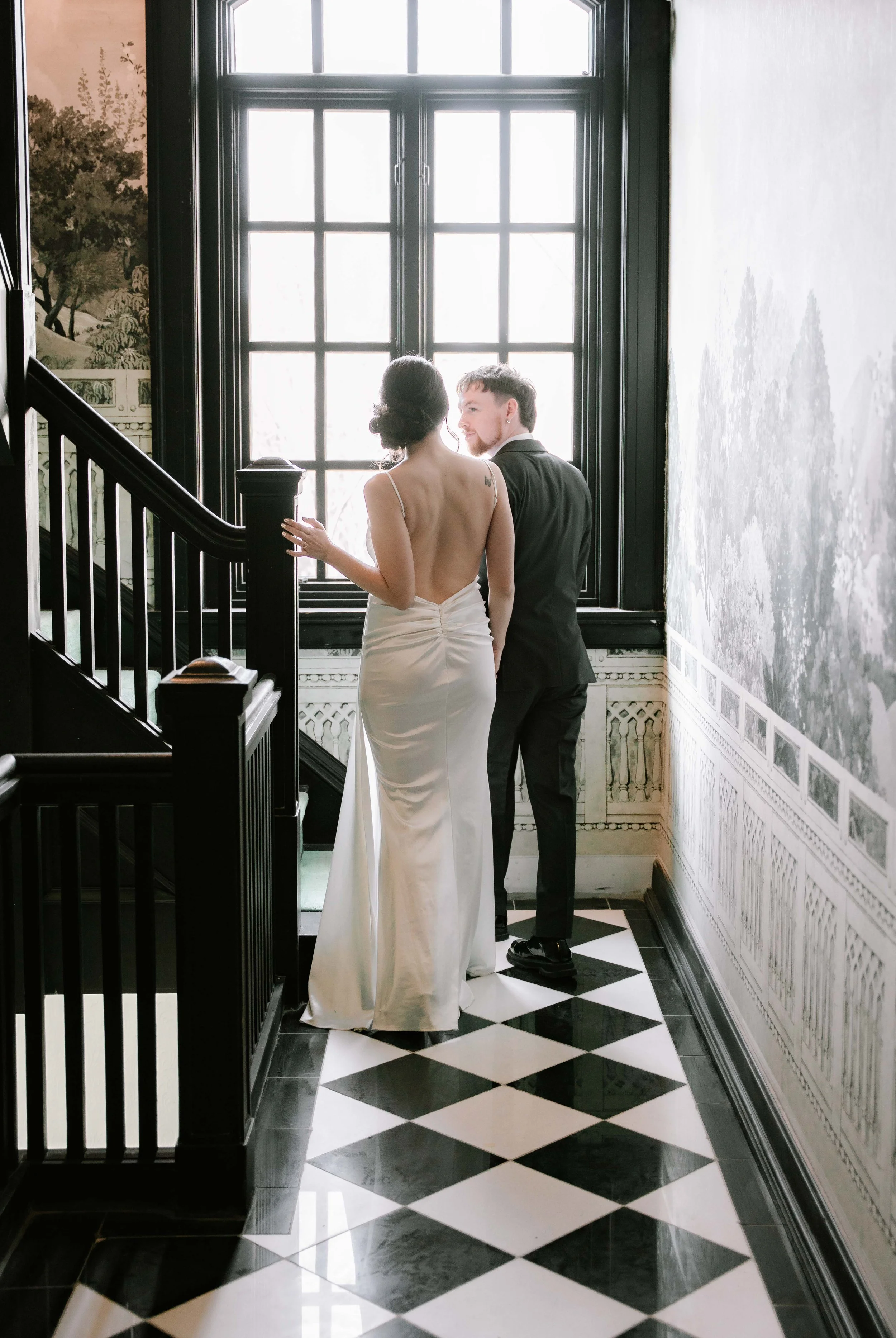 A couple standing on a staircase landing by a large window, the woman in a backless white dress and the man in a dark suit, looking at each other.