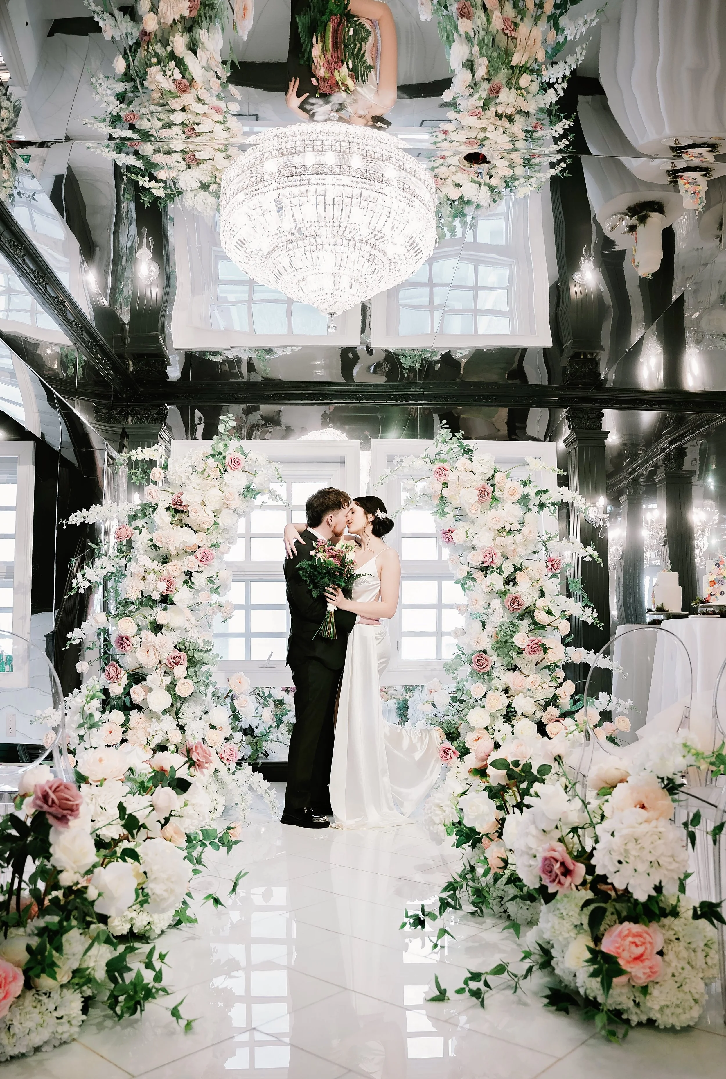 A bride and groom in wedding attire share a kiss in a decorated indoor wedding venue with floral arrangements and a large chandelier.